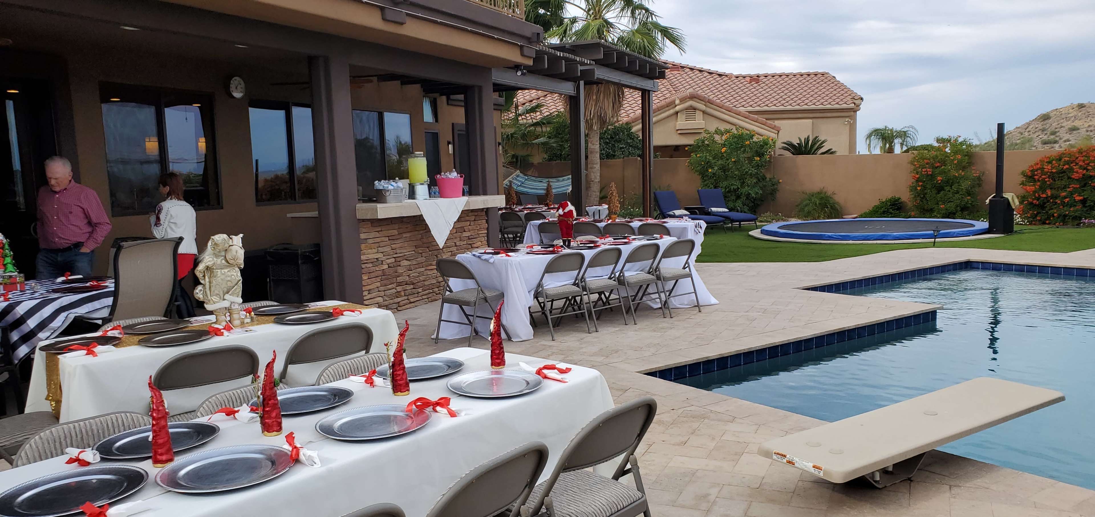The scene shows an outdoor dining setup with tables set for a meal beside a swimming pool in a residential backyard.