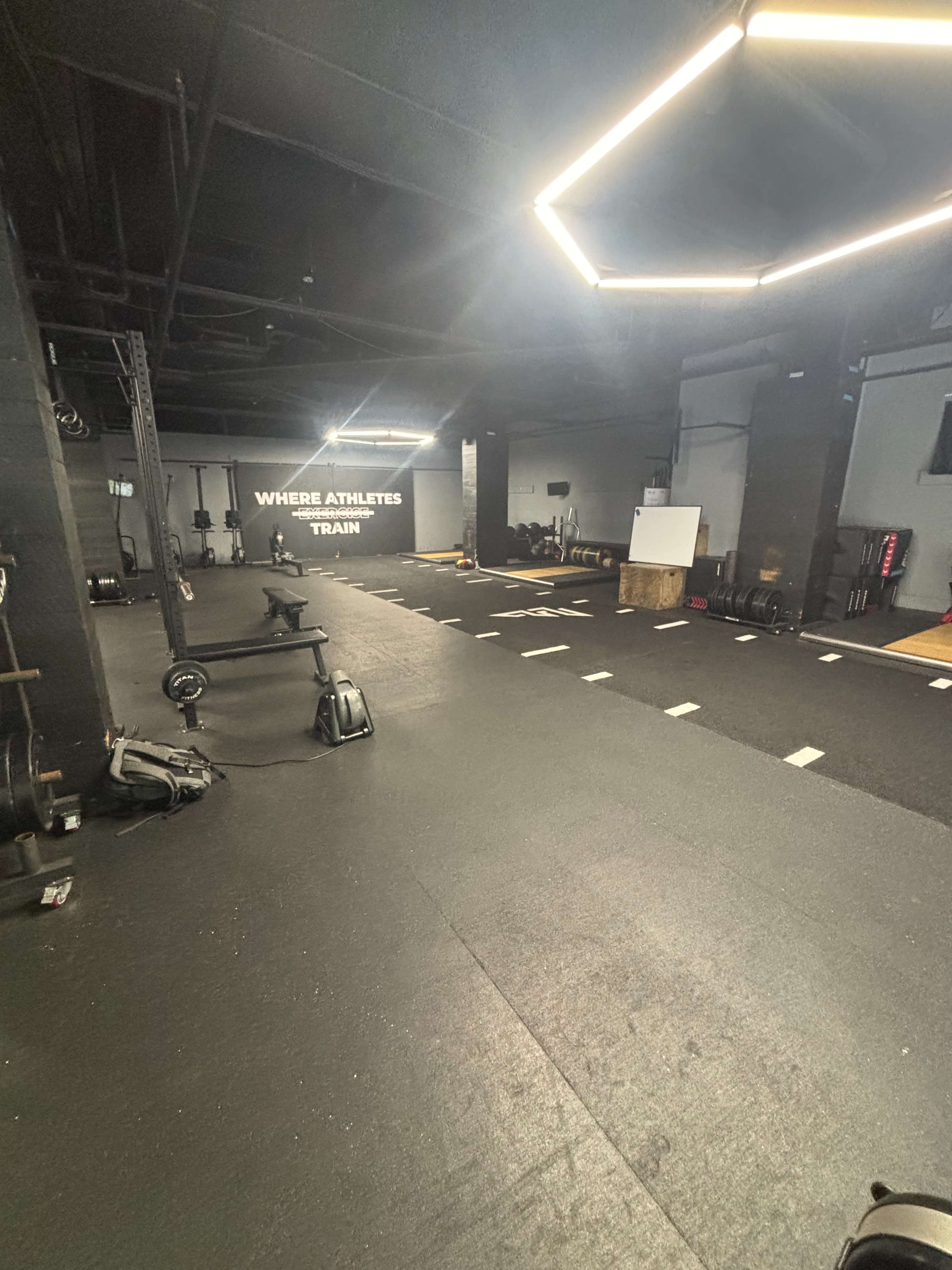 The image shows an empty gym space with black flooring, various fitness equipment, and a motivational wall that reads "WHERE ATHLETES TRAIN."