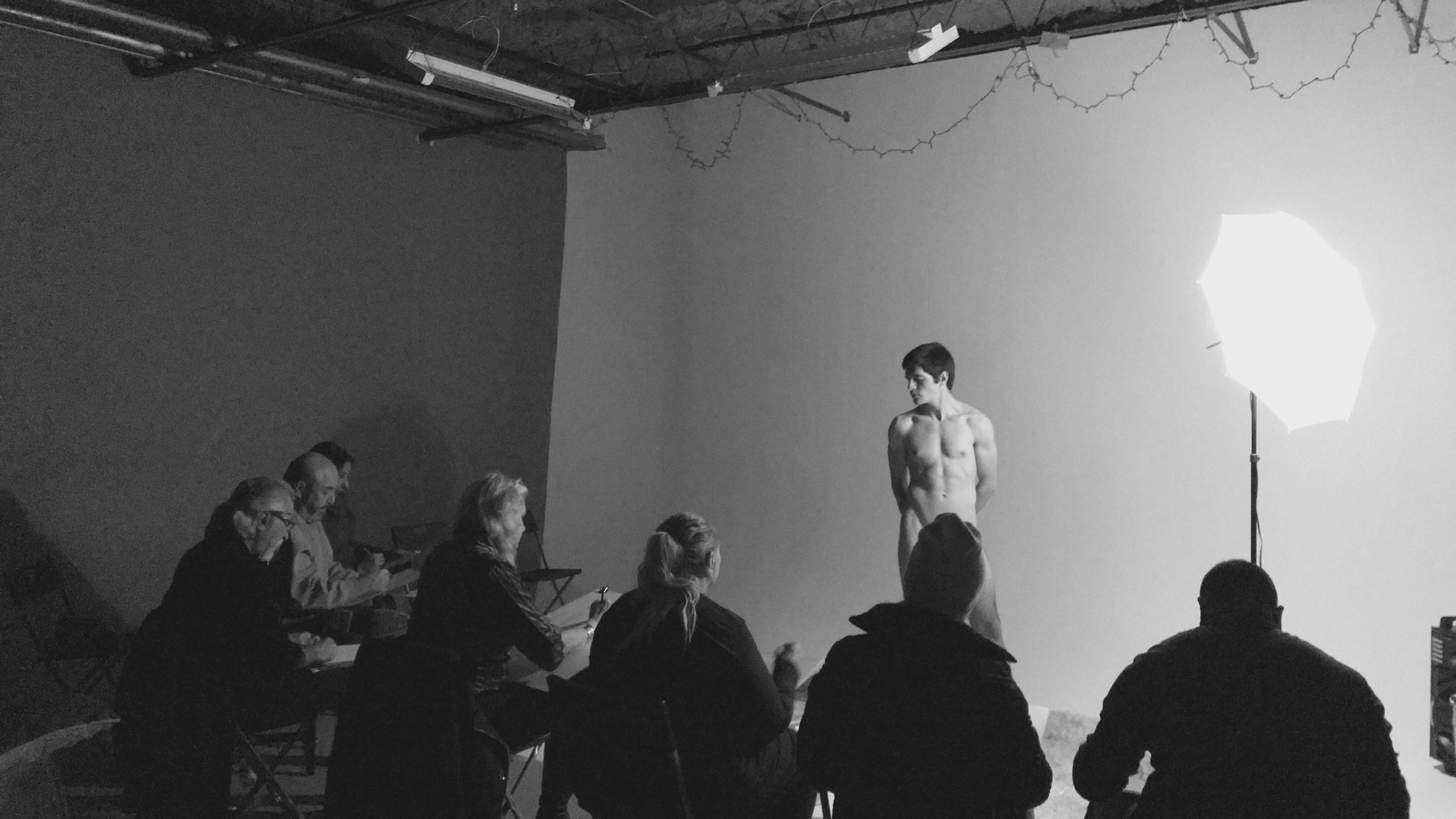 A model poses nude for a group of artists seated in chairs, with a bright light illuminating the scene.