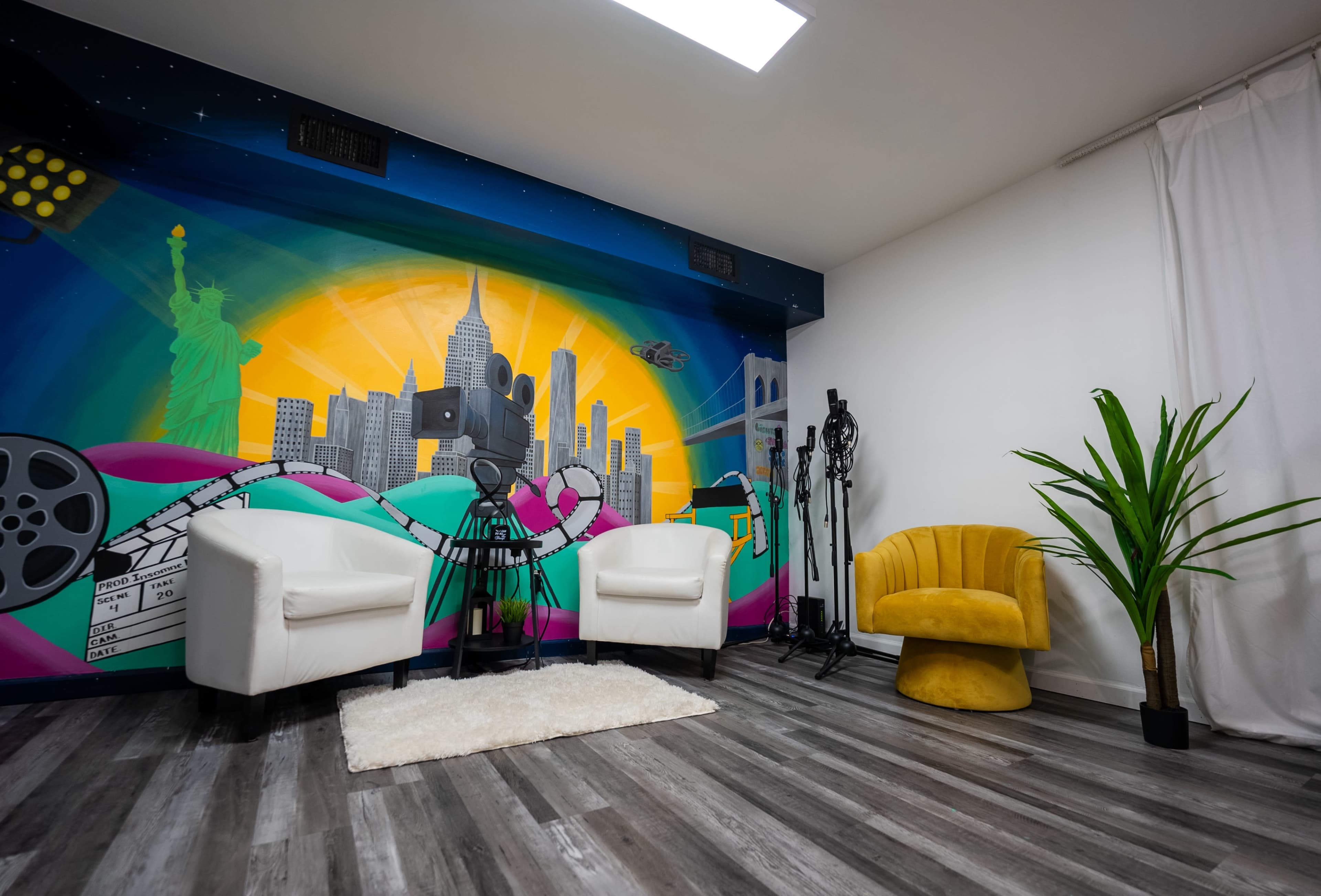 The image features a modern room with two white chairs, a yellow accent chair, a colorful mural of a city skyline, and a vibrant plant against a wooden floor.