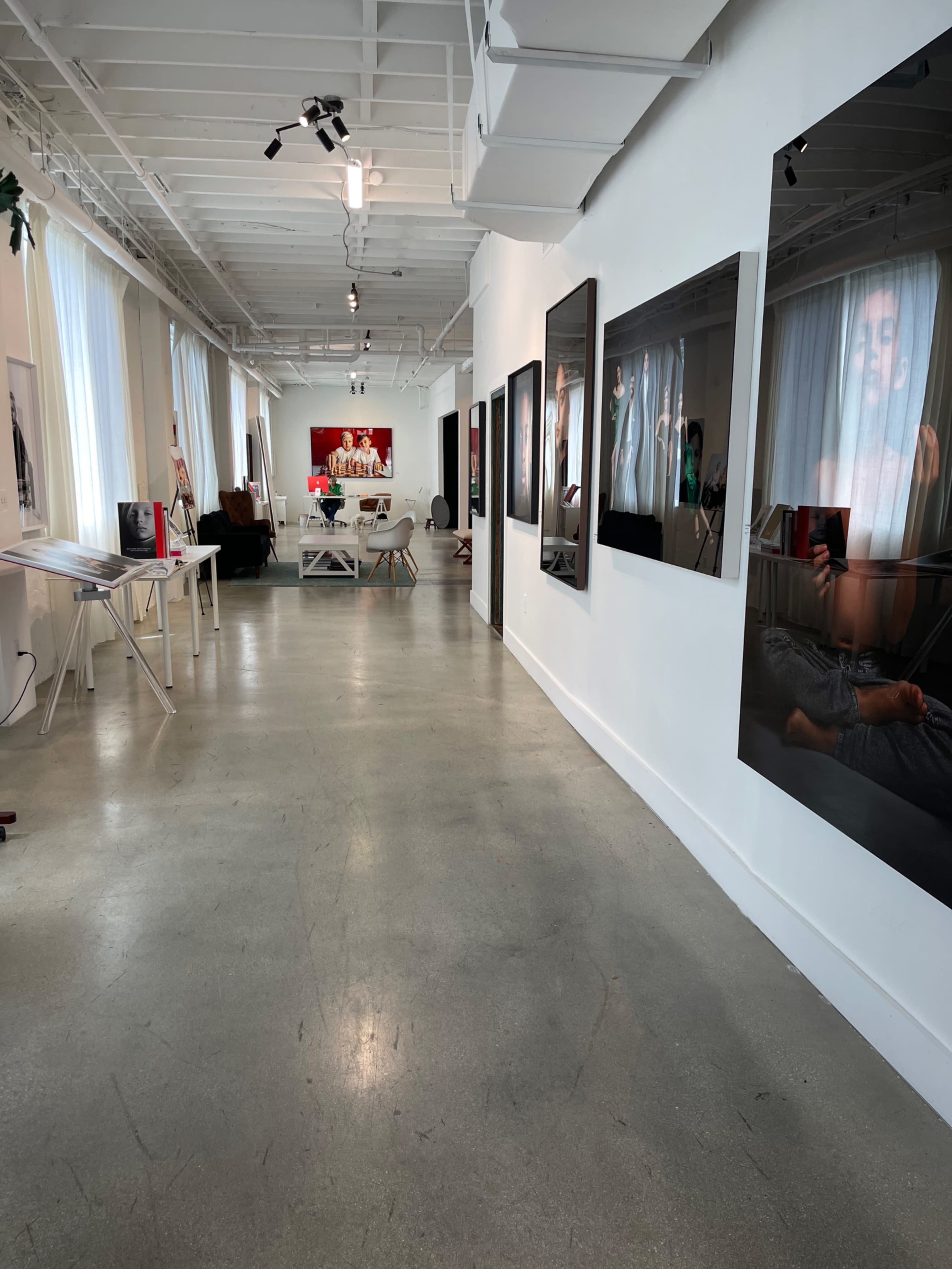 The image shows a modern art gallery with polished concrete floors, white walls displaying various artworks, and large windows providing ample natural light.
