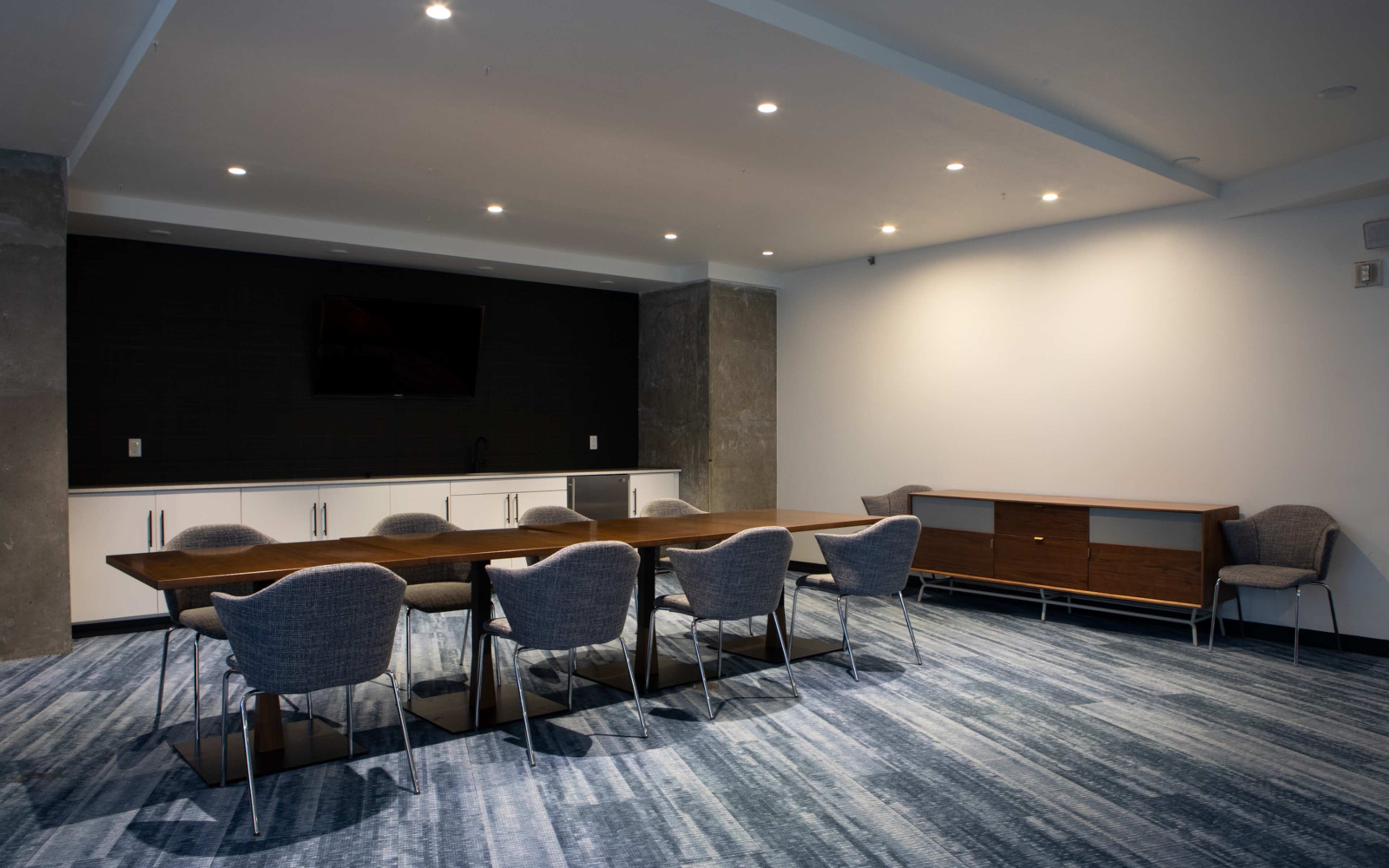 A modern conference room with a long wooden table surrounded by gray chairs, and a wall-mounted television on a dark background.