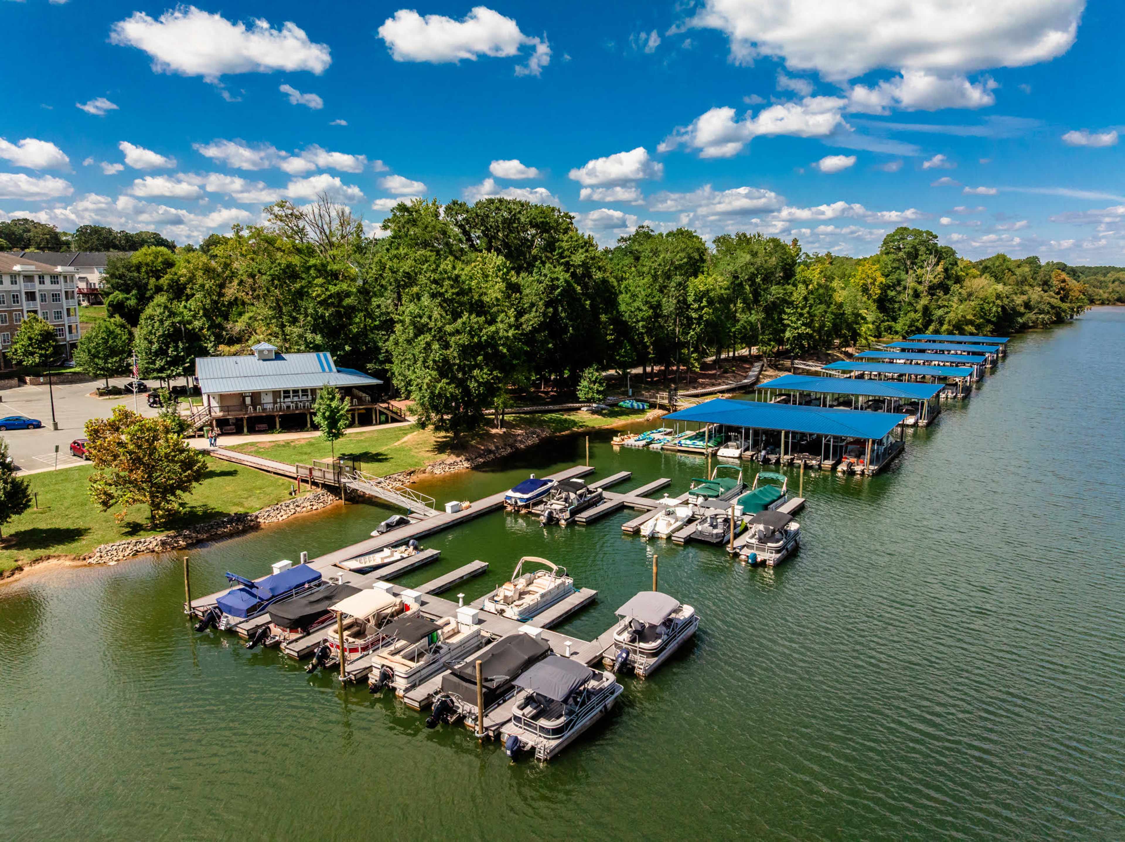 The image shows a marina with several boats docked along the water, surrounded by green trees and a building in the background.