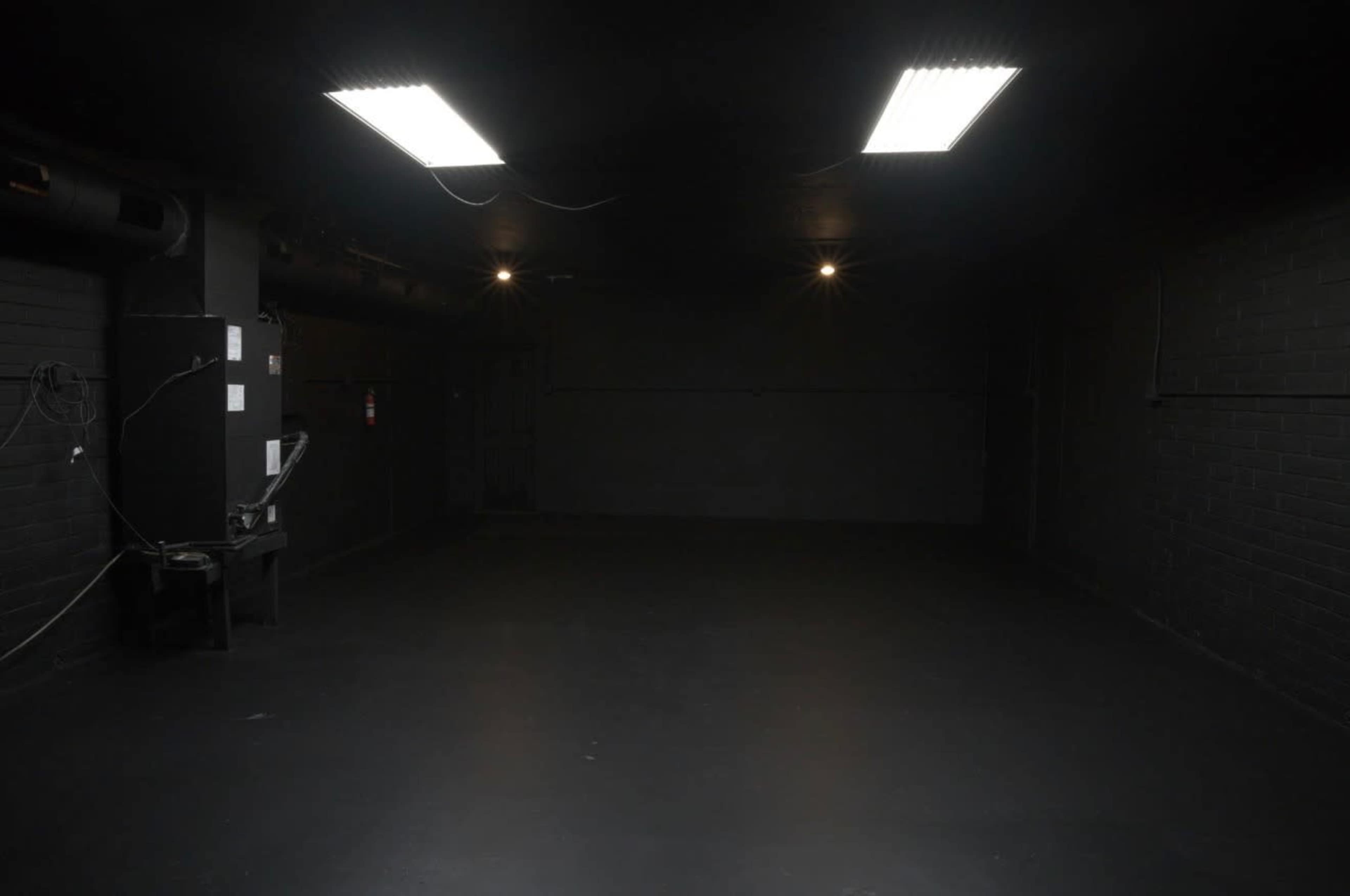 The image shows a dimly lit, empty room with black walls and a smooth black floor, featuring overhead fluorescent lights and a small machine in one corner.