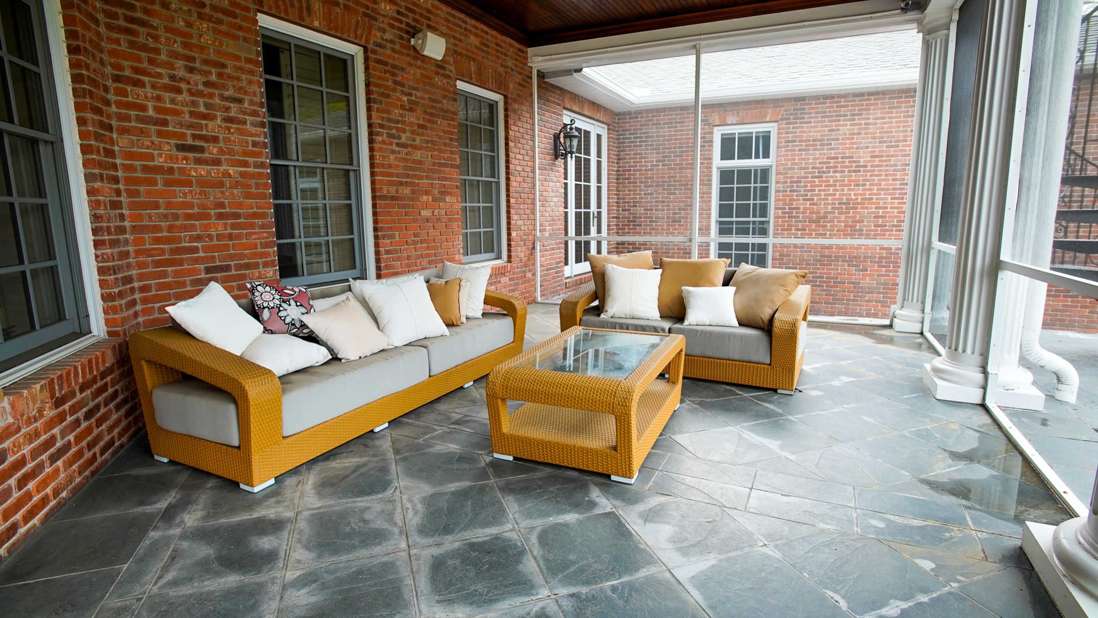 A screened-in porch featuring a wicker sofa with cushions, a glass coffee table, and a stone floor, surrounded by brick walls and large windows.