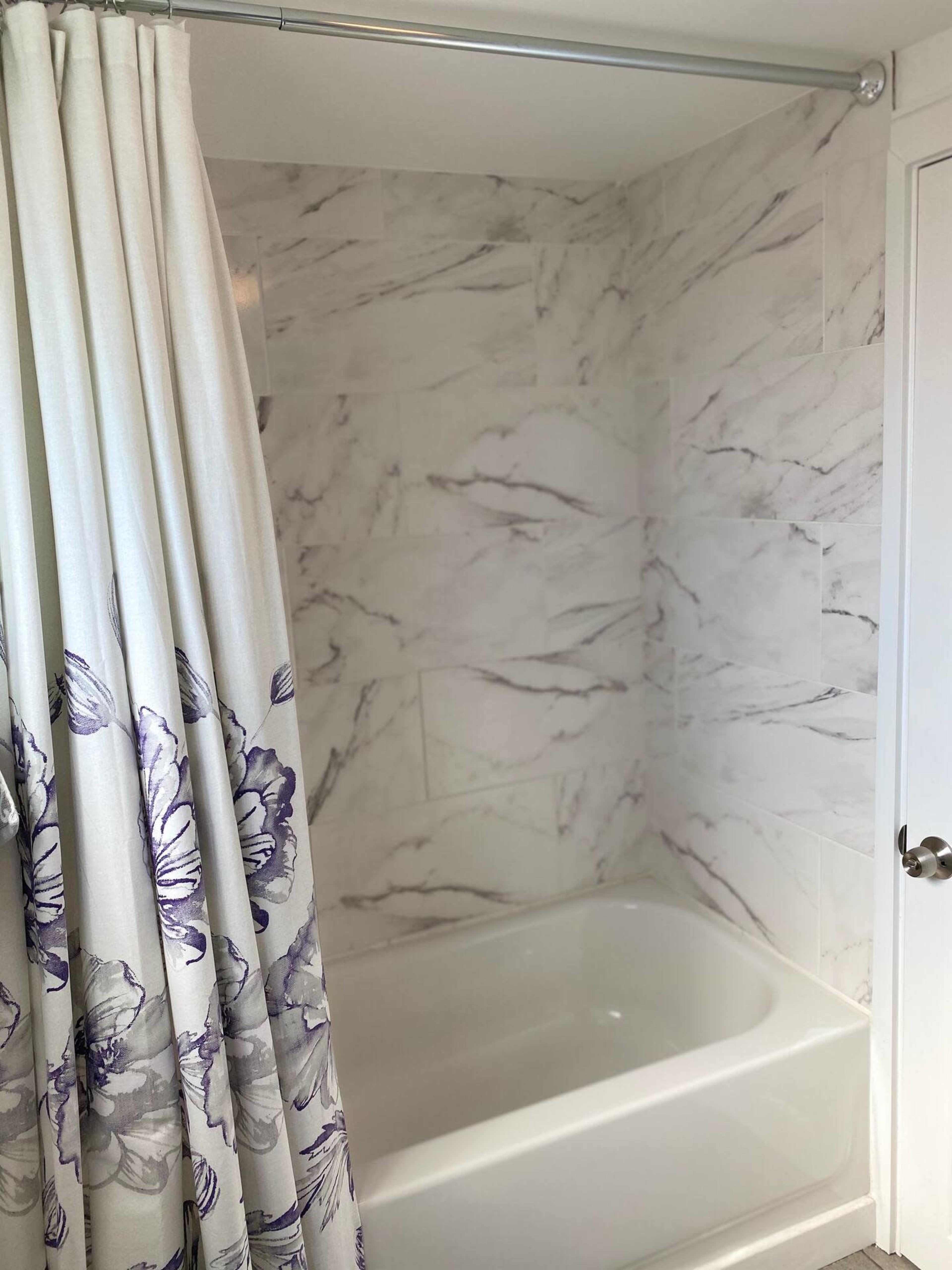 The image shows a bathroom with a shower area featuring a white tub and a floral shower curtain against marble-patterned walls.