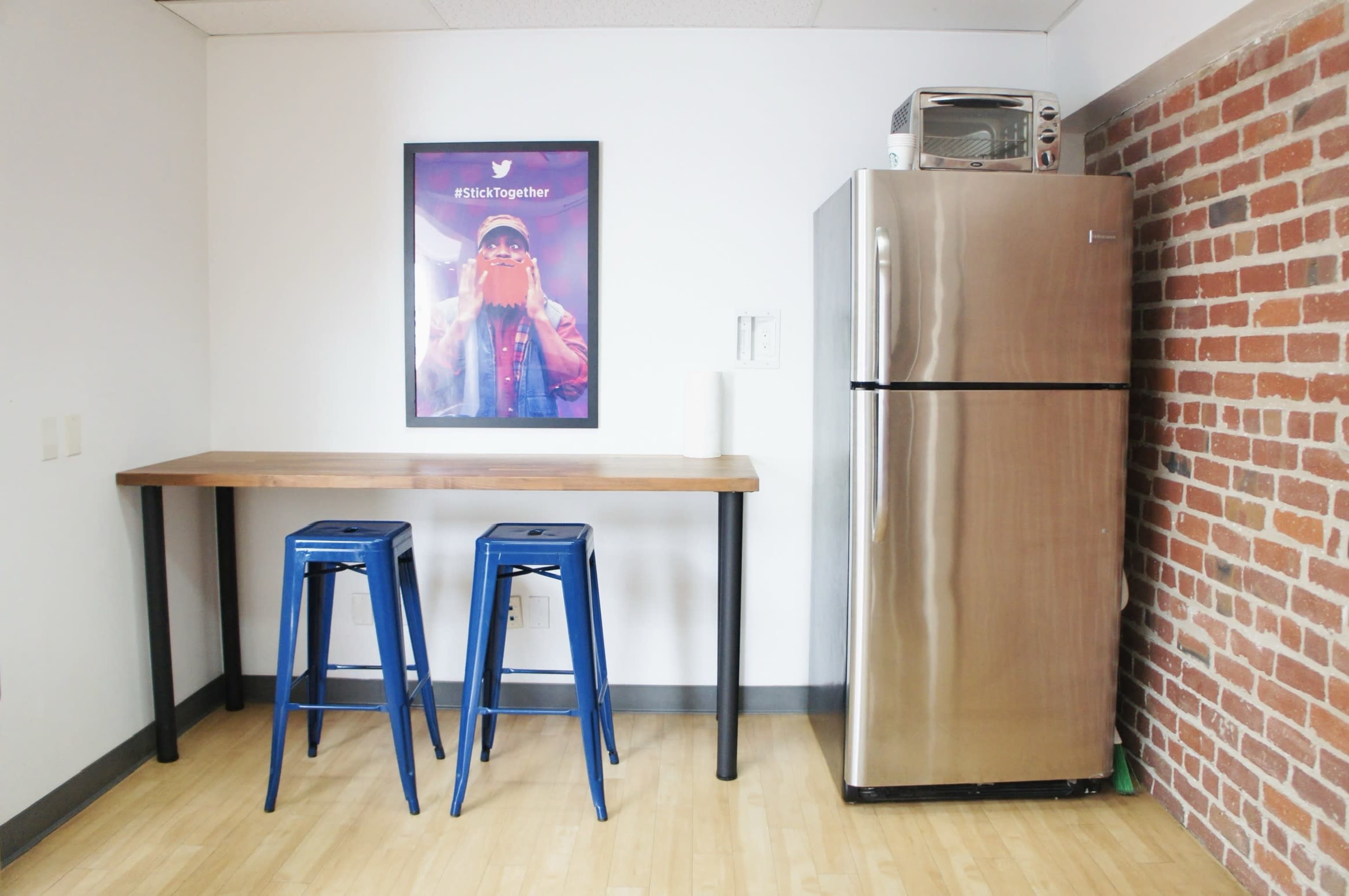 A small kitchen area features a metallic refrigerator, a microwave above it, a wooden table with two blue stools, and a framed poster on the wall.