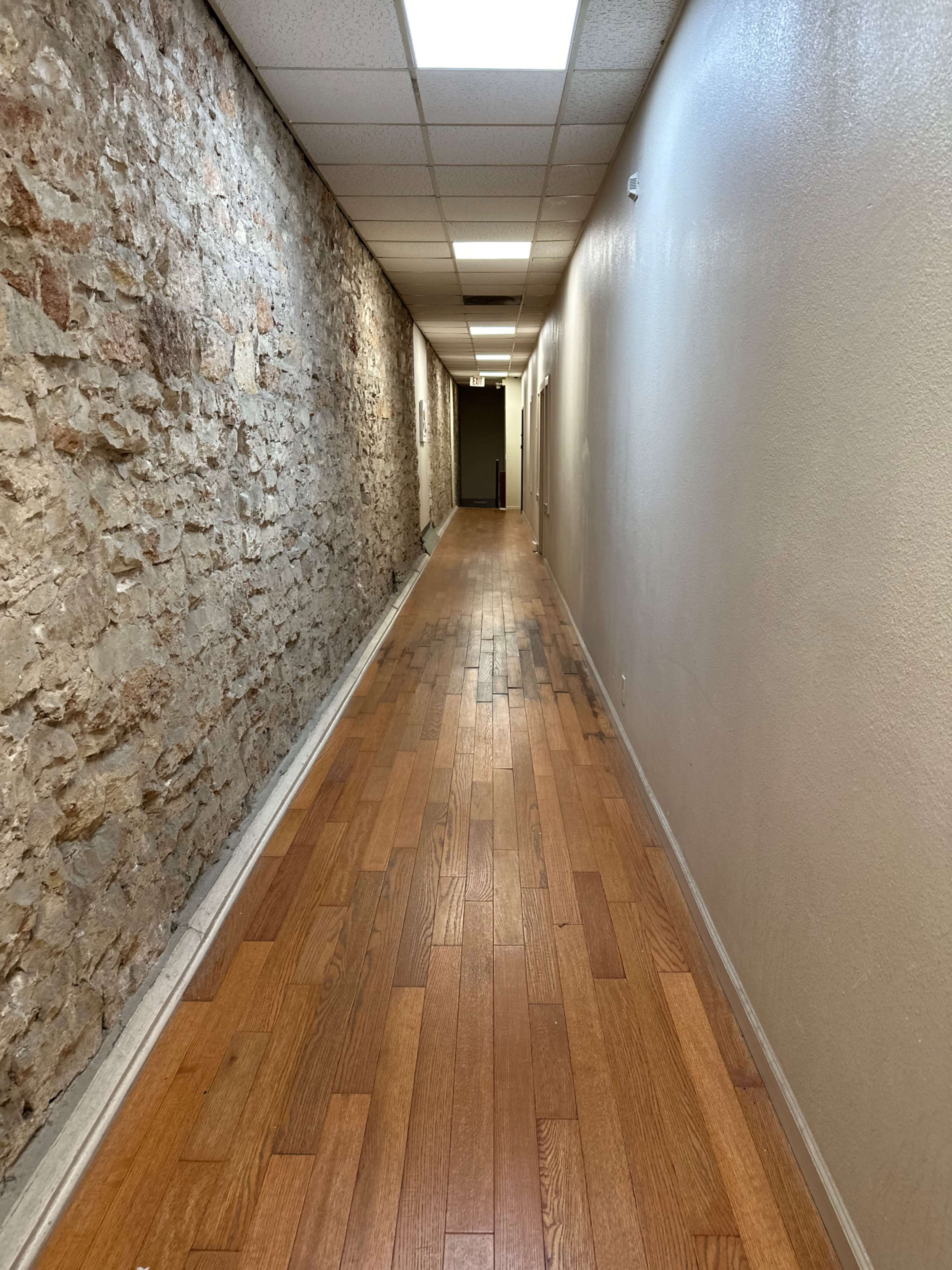 The image shows a long hallway lined with a stone wall on one side and wooden flooring leading to a distant door.