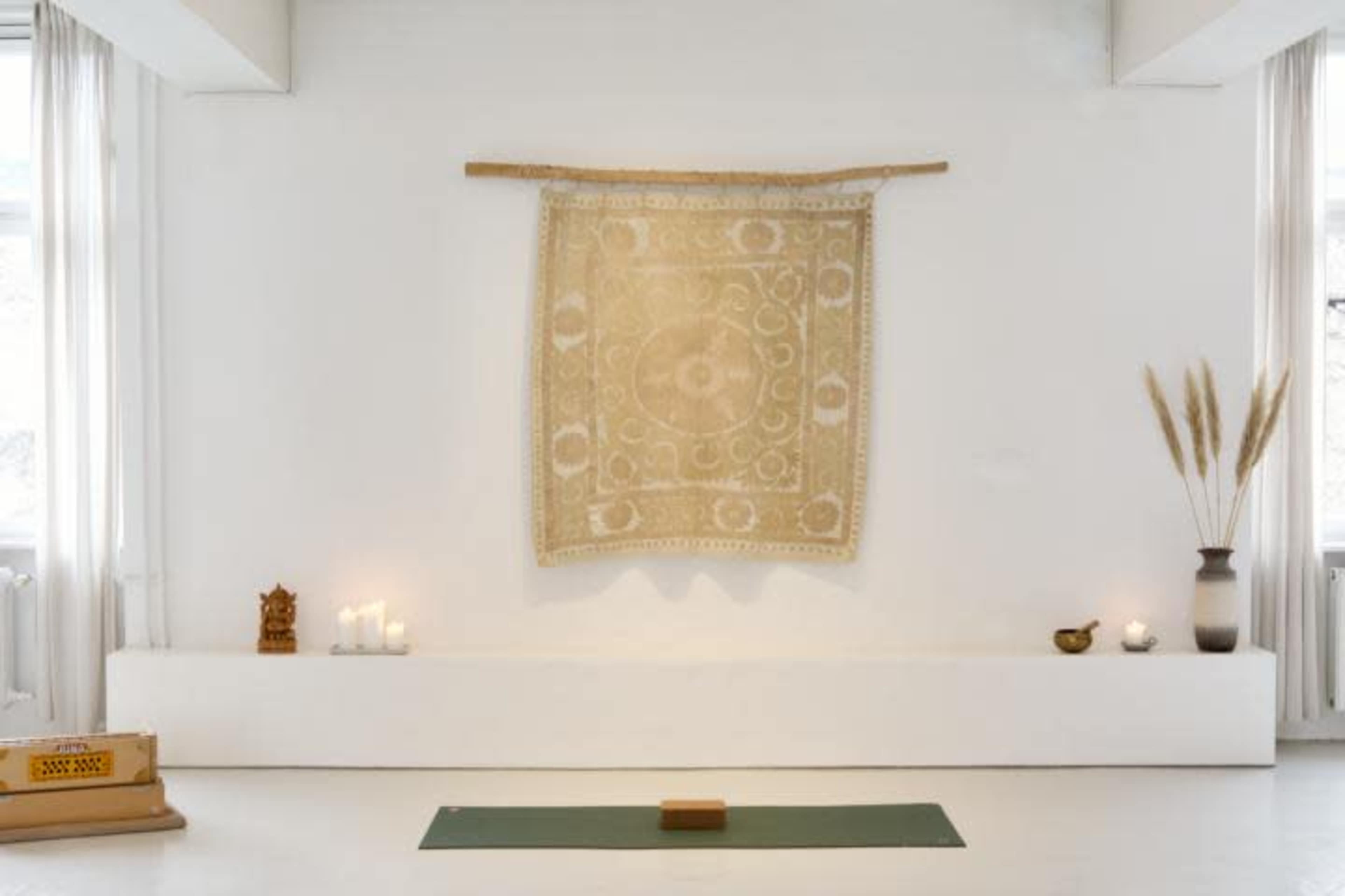 A minimalistic room with a green yoga mat on the floor, a tapestry hanging on the wall, candles on a ledge, and decorative elements including a wooden figure and a bowl.