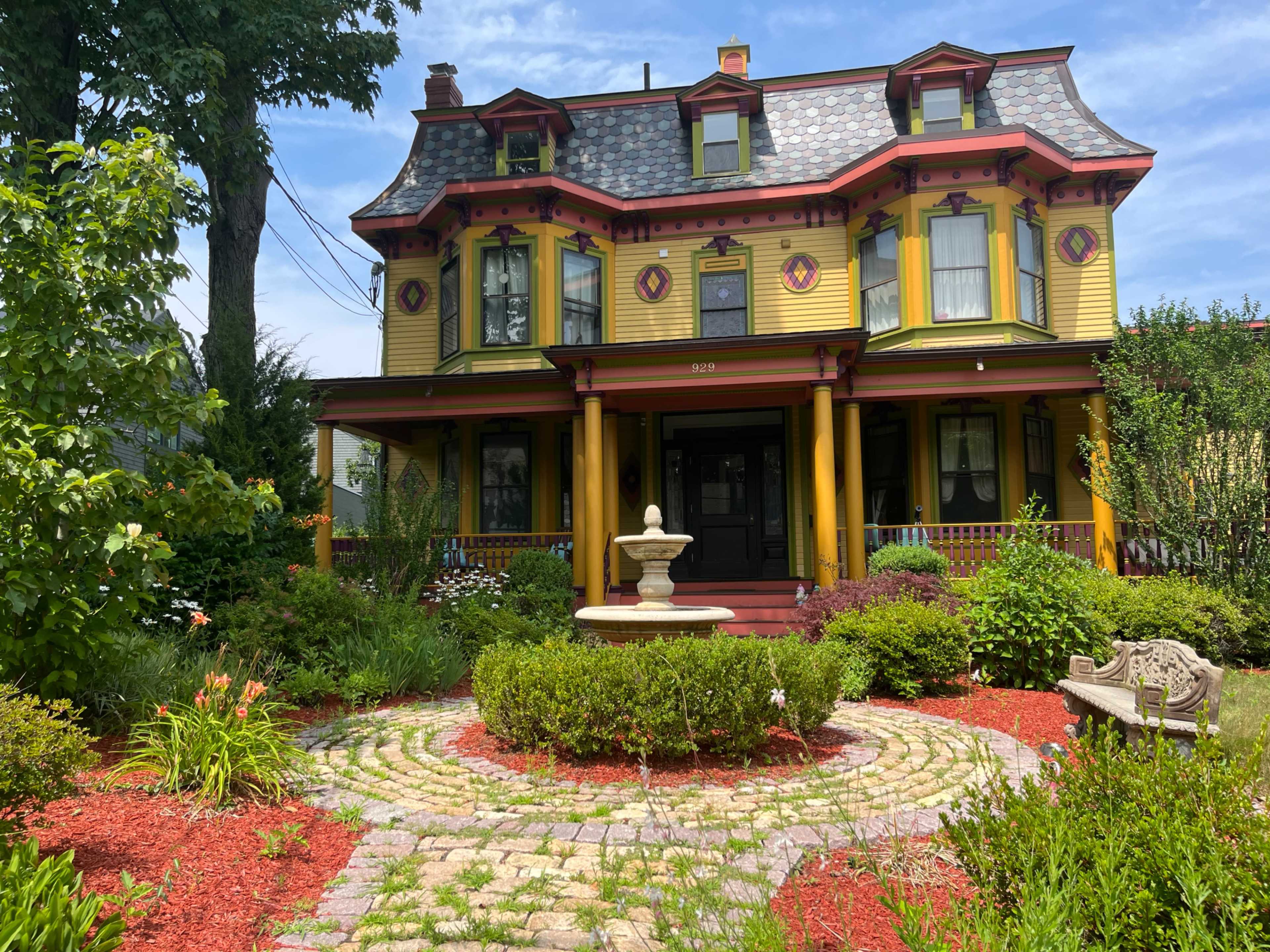 A Victorian-style house with yellow siding and red accents stands prominently in a landscaped garden featuring a stone fountain and circular stone pathway.