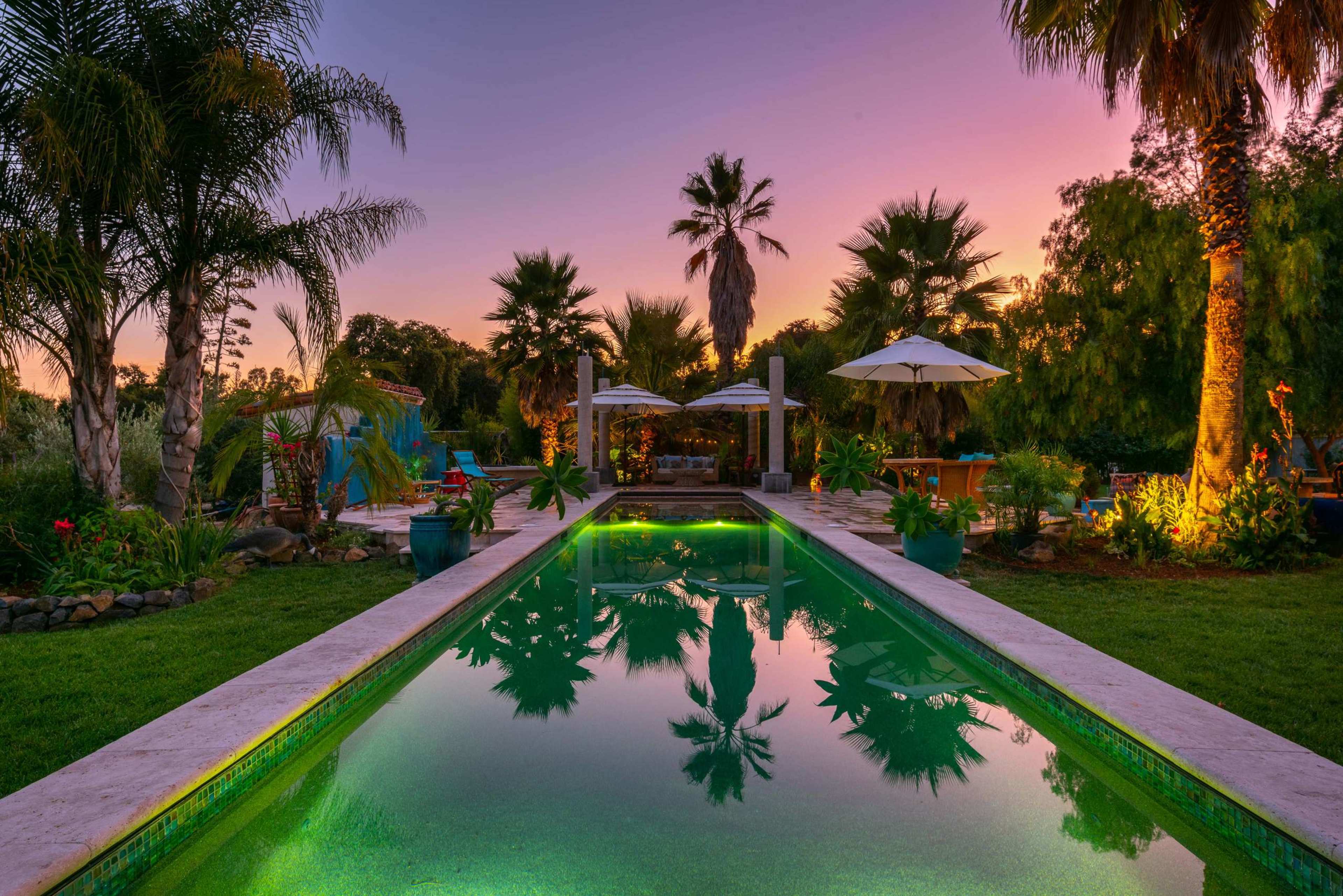 A tranquil garden features a swimming pool reflecting palm trees and the colorful sky at dusk.