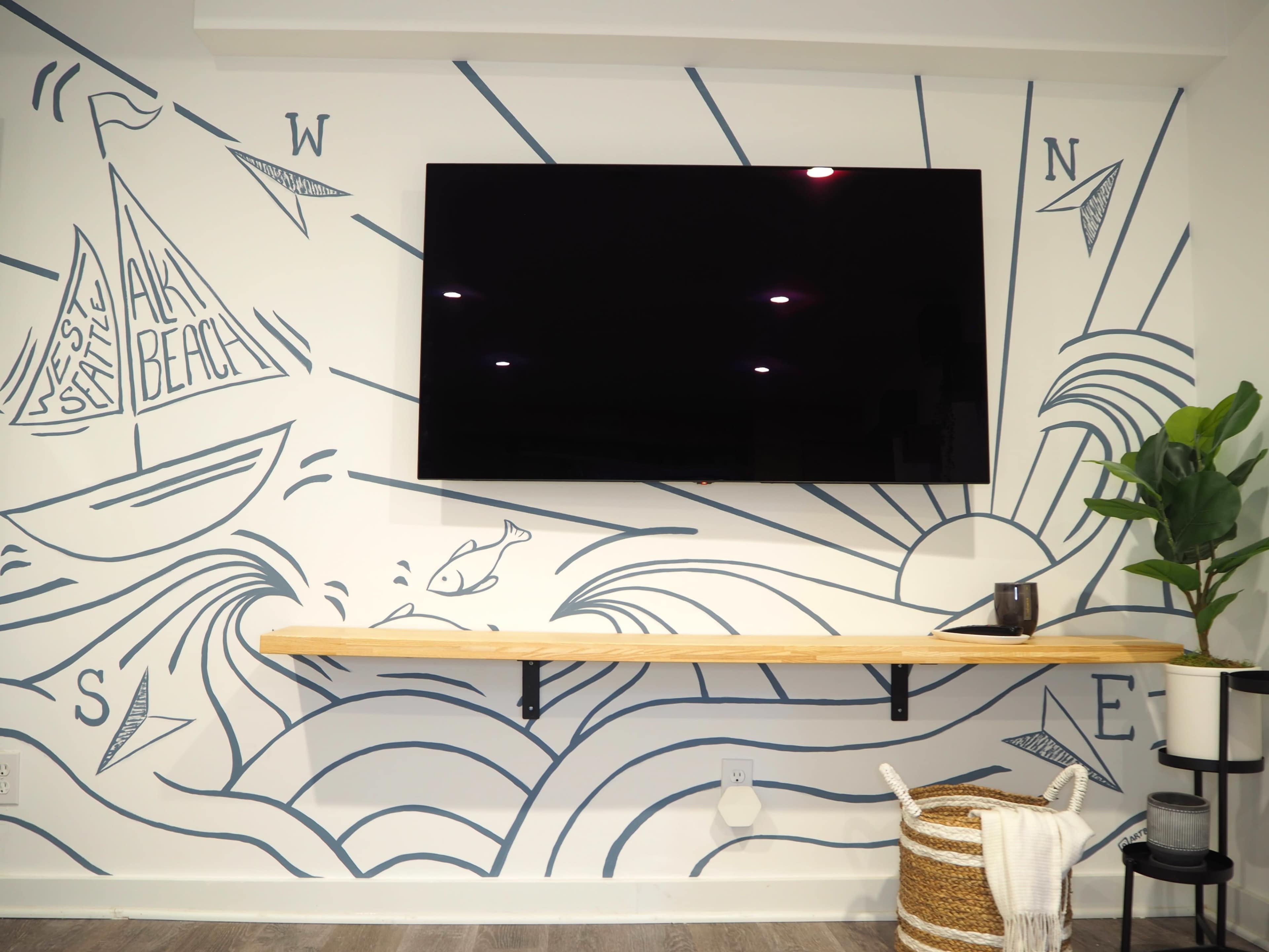 A wall features a large, flat-screen TV above a wooden shelf with a decorative plant, surrounded by a mural depicting waves, boats, and a sunburst design.