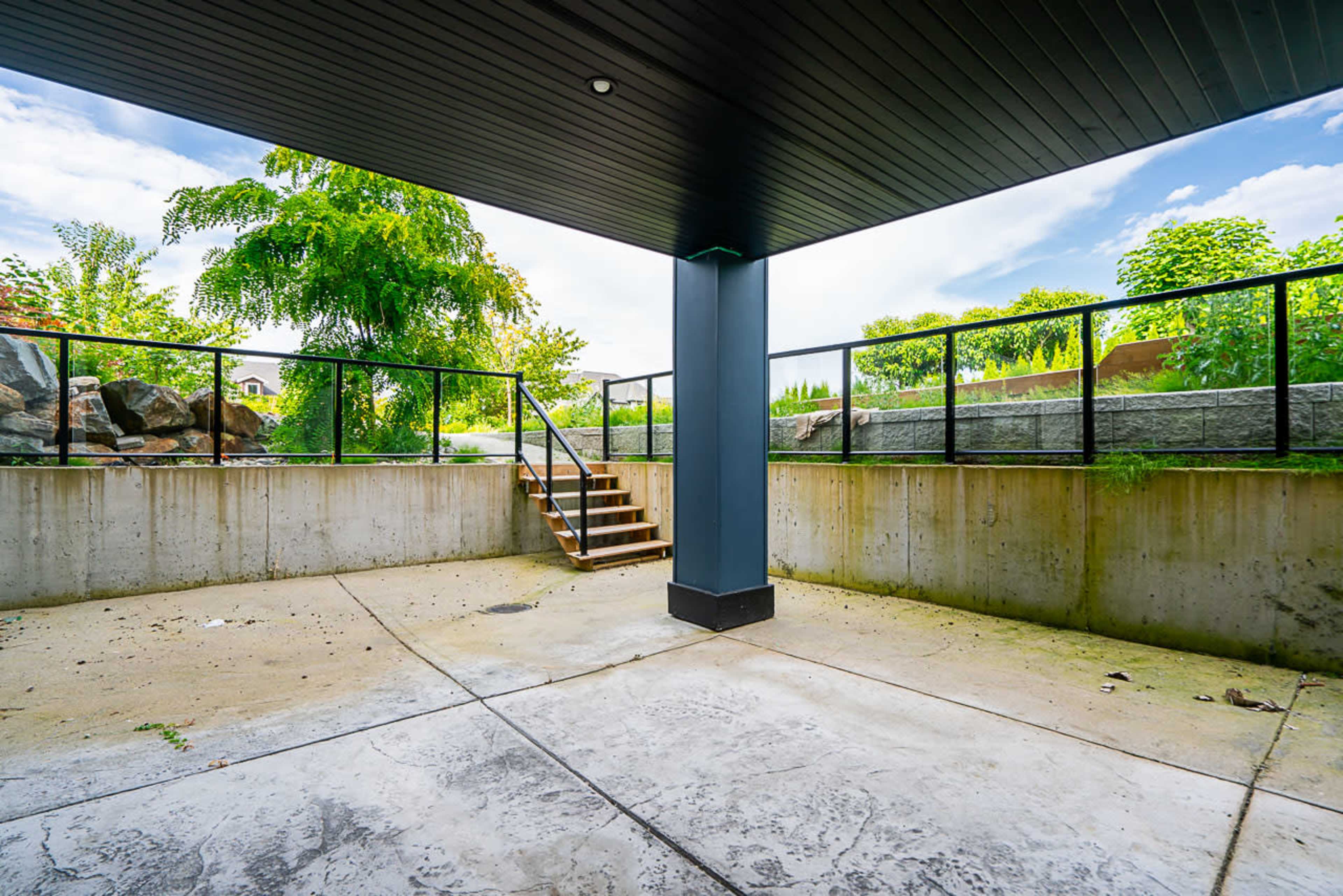 A concrete outdoor space enclosed by a black railing, with a staircase leading up and green foliage visible in the background under a blue sky.