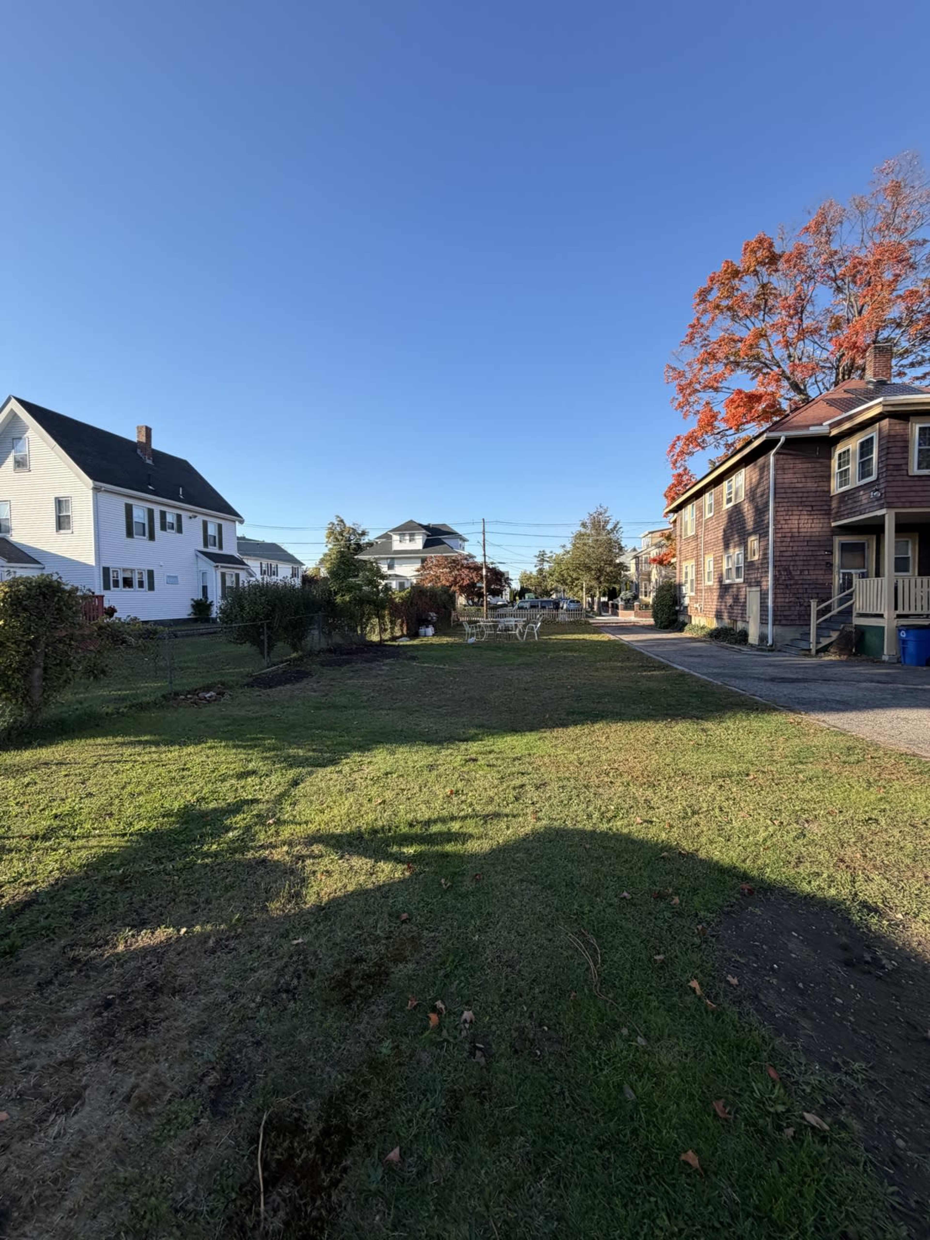 The scene shows a grassy area between houses, with a clear blue sky and a few autumn trees in the background.