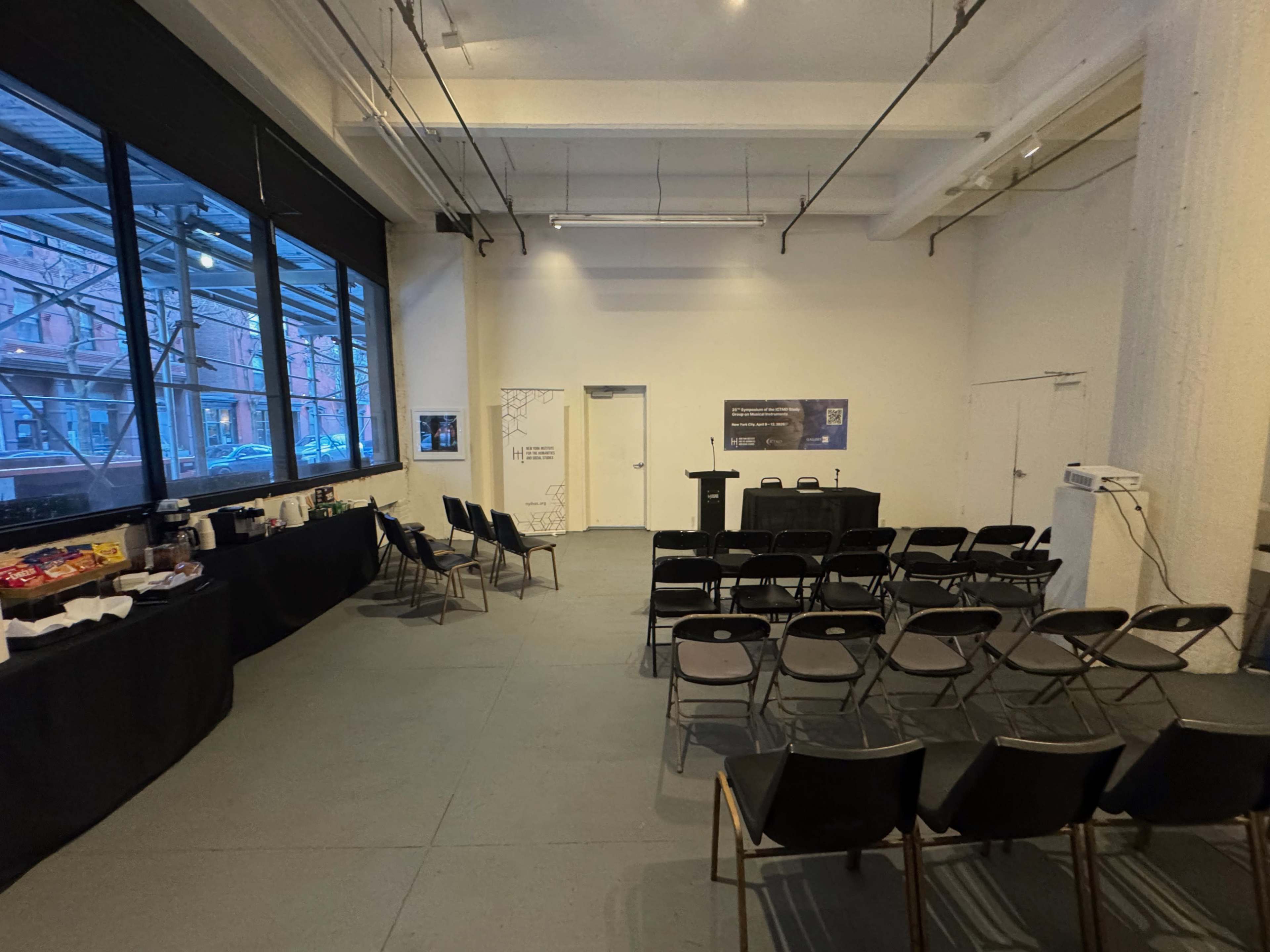 The image shows a spacious, empty event room with rows of chairs set up in front of a podium, accompanied by a long table displaying snacks and beverages along one side.