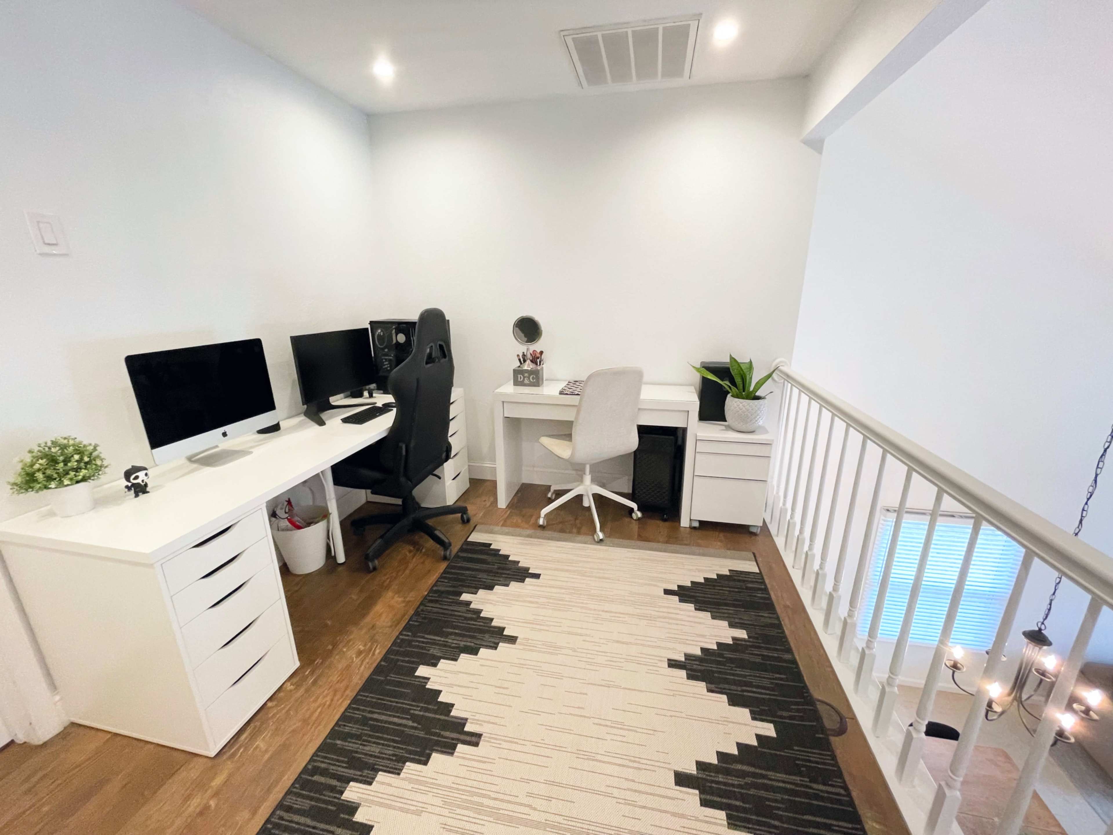 The image shows a modern home office setup with two desks, an ergonomic chair, a computer, and a decorative plant near a staircase.