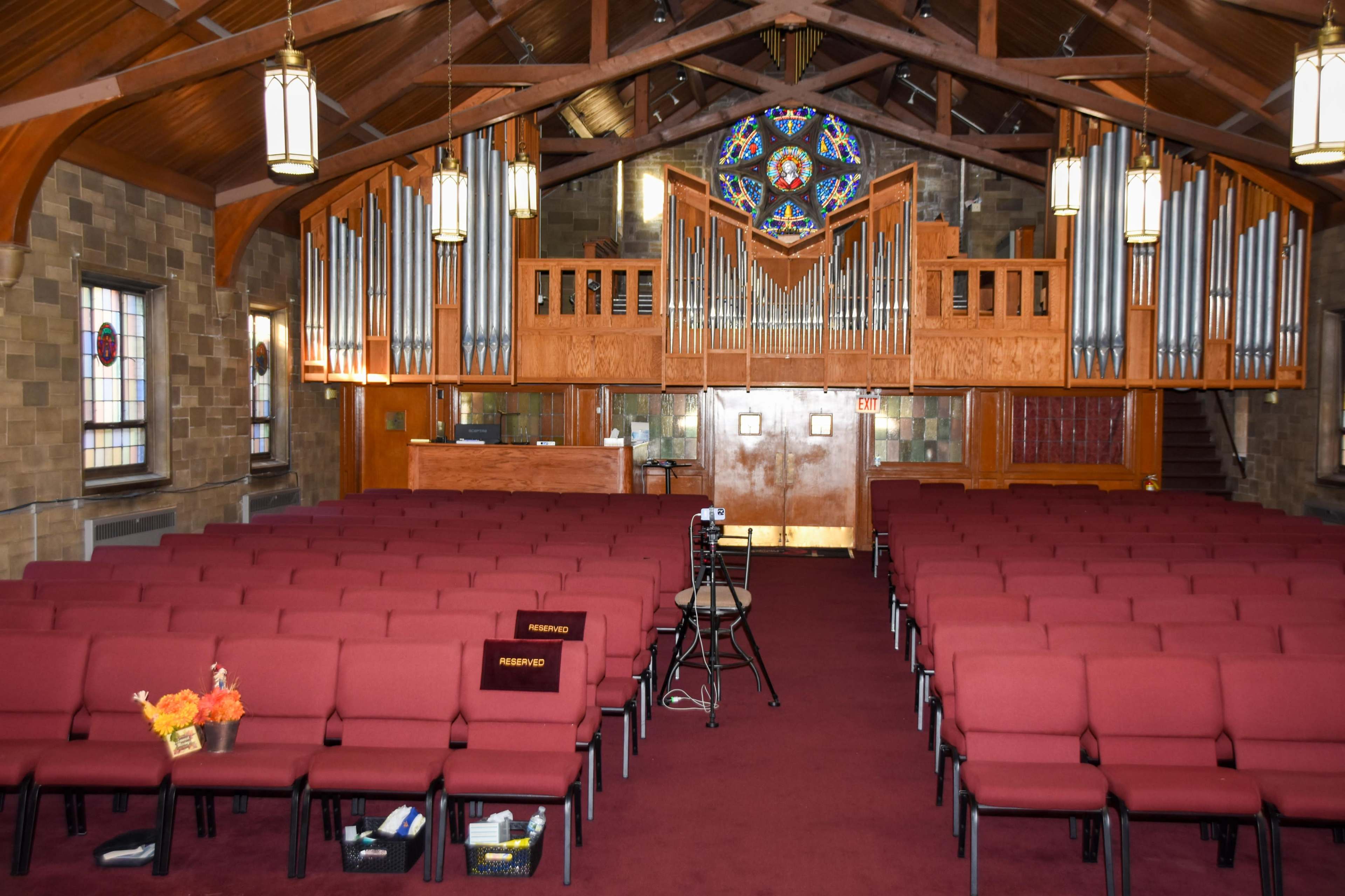 The image shows the interior of a church with rows of red upholstered chairs facing a wooden organ and a stained glass window in the background.