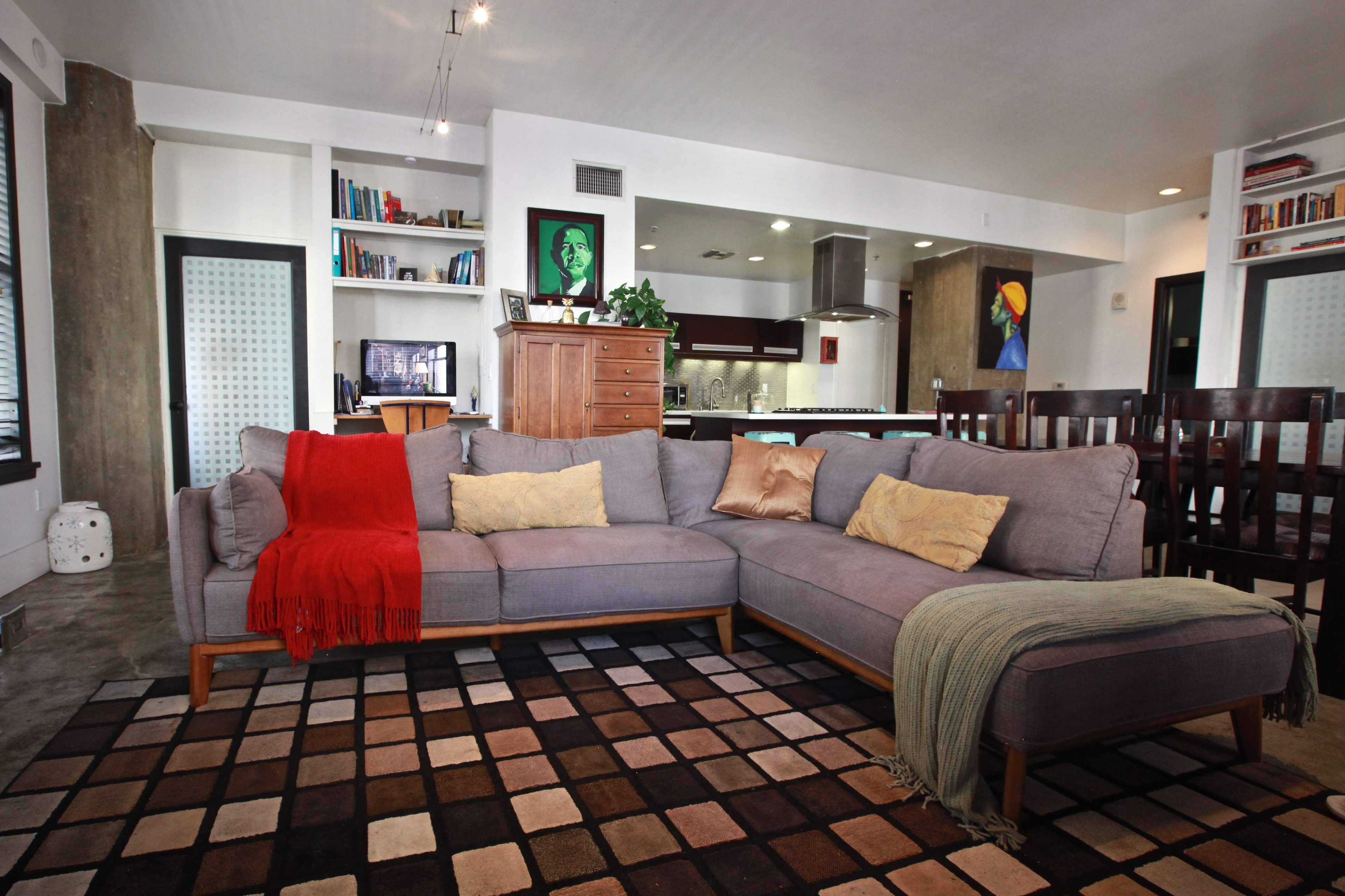 The image shows a contemporary living room featuring a grey sectional sofa with a red throw, a patterned area rug, and a kitchen visible in the background.