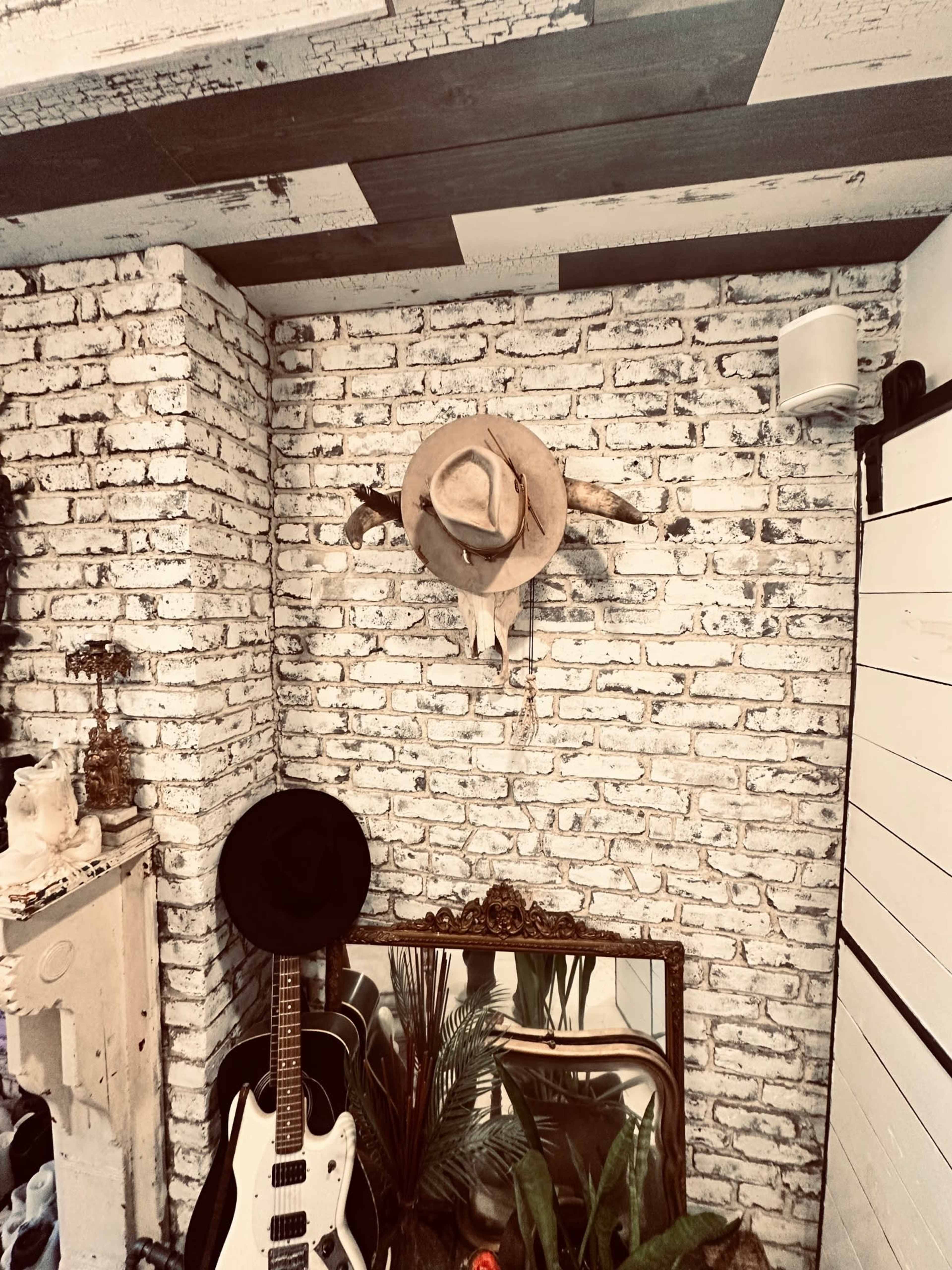 The image shows a corner of a room featuring a brick wall, a mounted pair of horns, a hat hanging above, a guitar leaning against the wall, and a vintage mirror reflecting a plant.