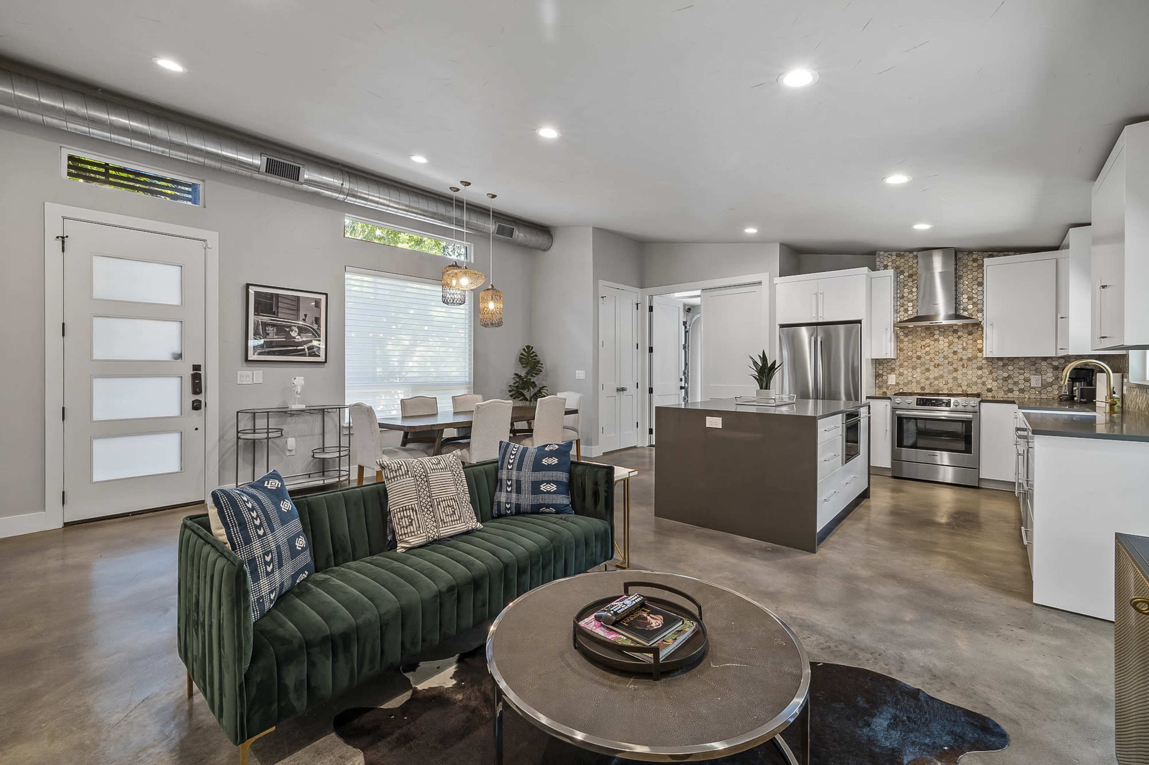 The image shows a modern open-concept living space featuring a green velvet sofa, a round coffee table, and a kitchen with stainless steel appliances and a large island.
