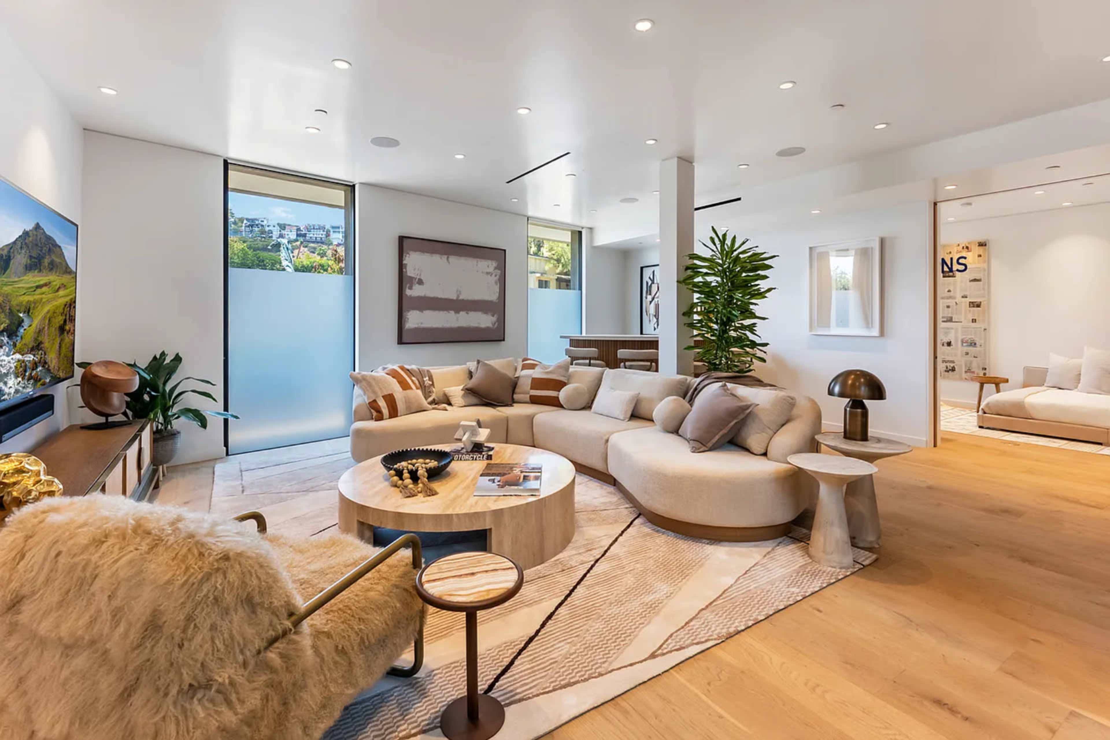 The image shows a modern living room with a curved sofa, a round coffee table, and a large, textured armchair, surrounded by bright, natural light from windows.