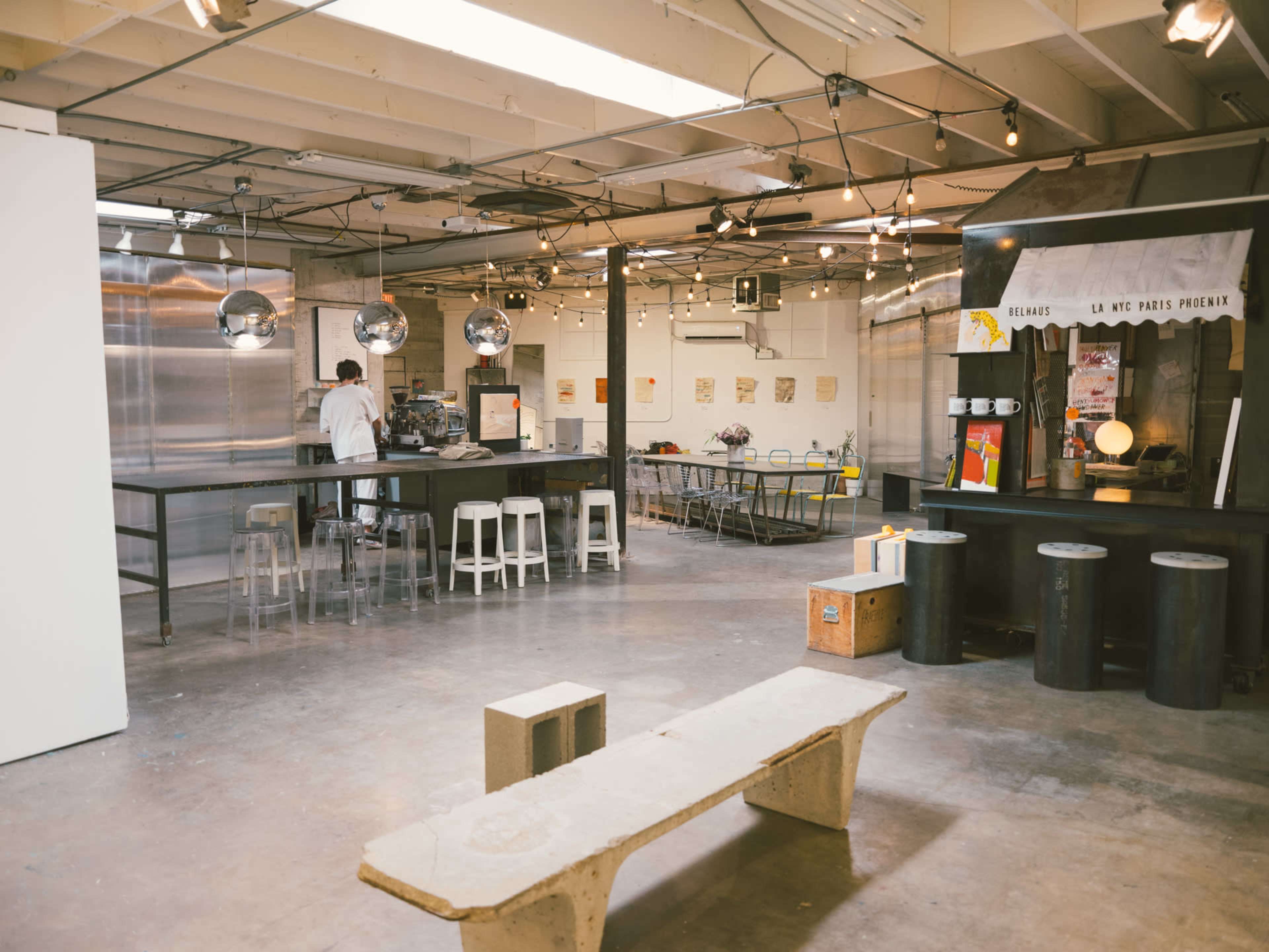The image shows a spacious industrial kitchen and dining area with metal fixtures, a long table, and stools, illuminated by hanging bulbs.