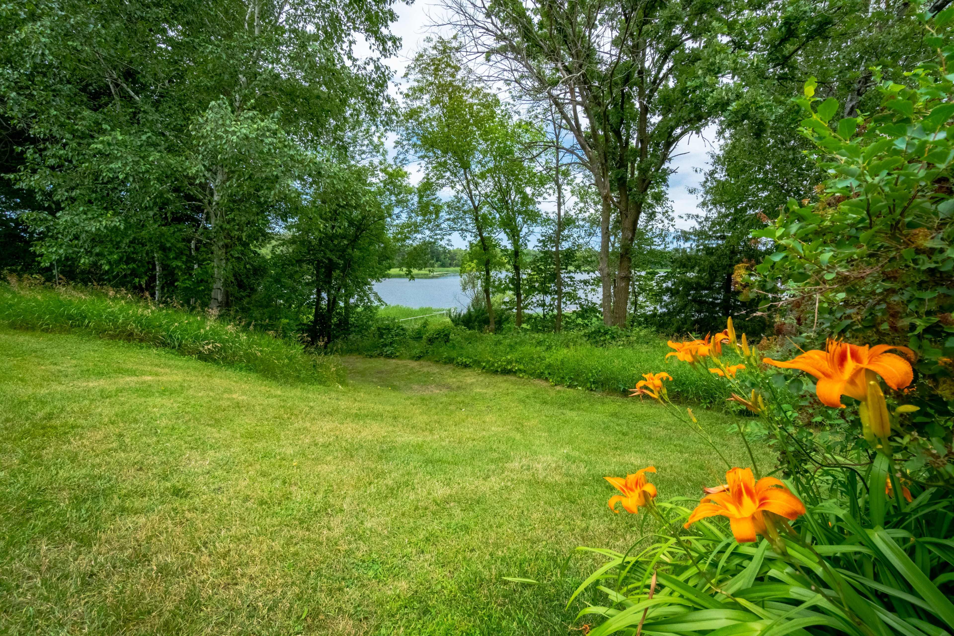 A vibrant garden with orange flowers leads to a grassy area that overlooks a lake surrounded by trees.