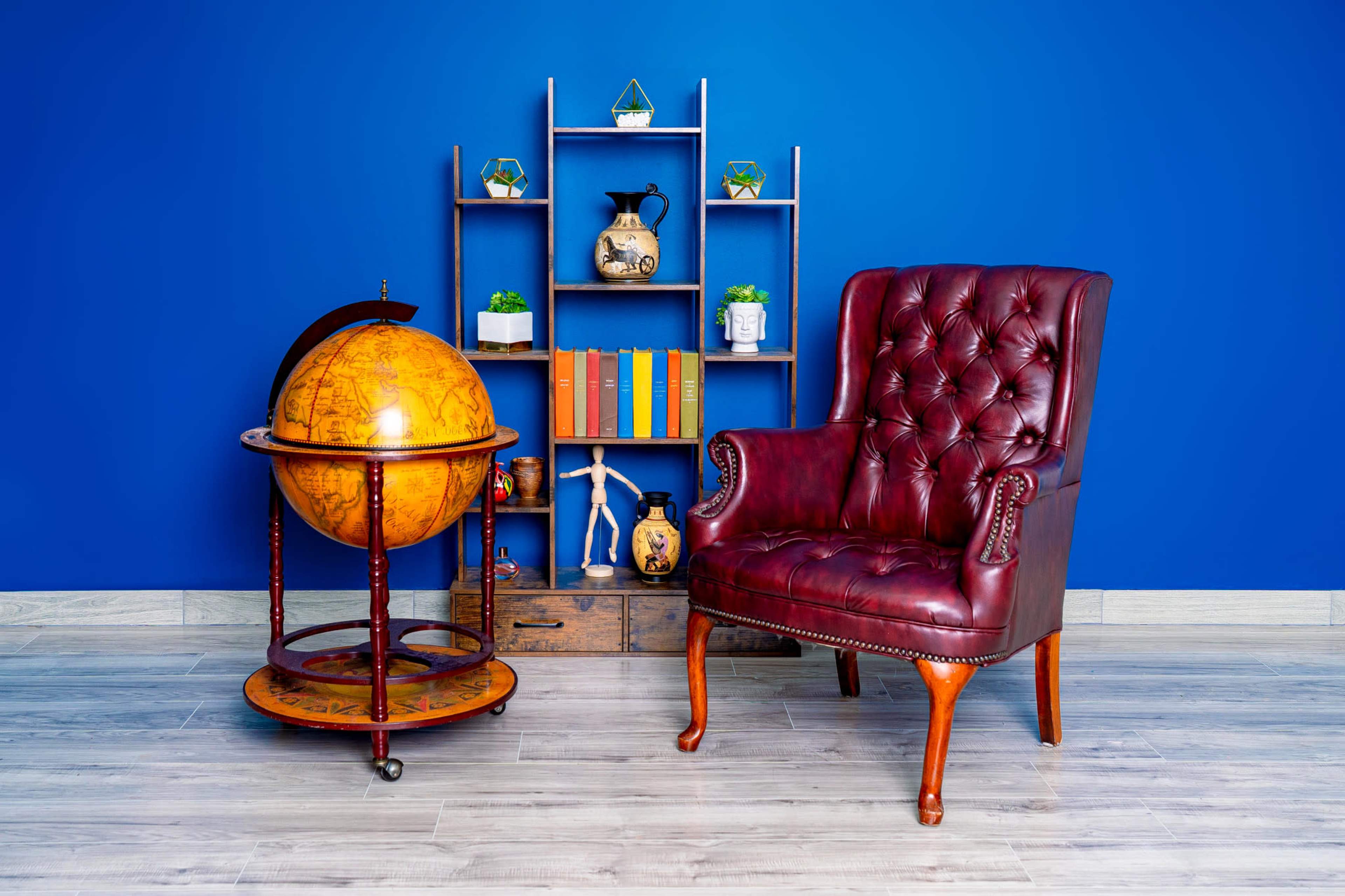 The image features a red leather, tufted armchair next to a wooden globe on a stand, with a blue wall in the background and a shelf displaying plants and books.