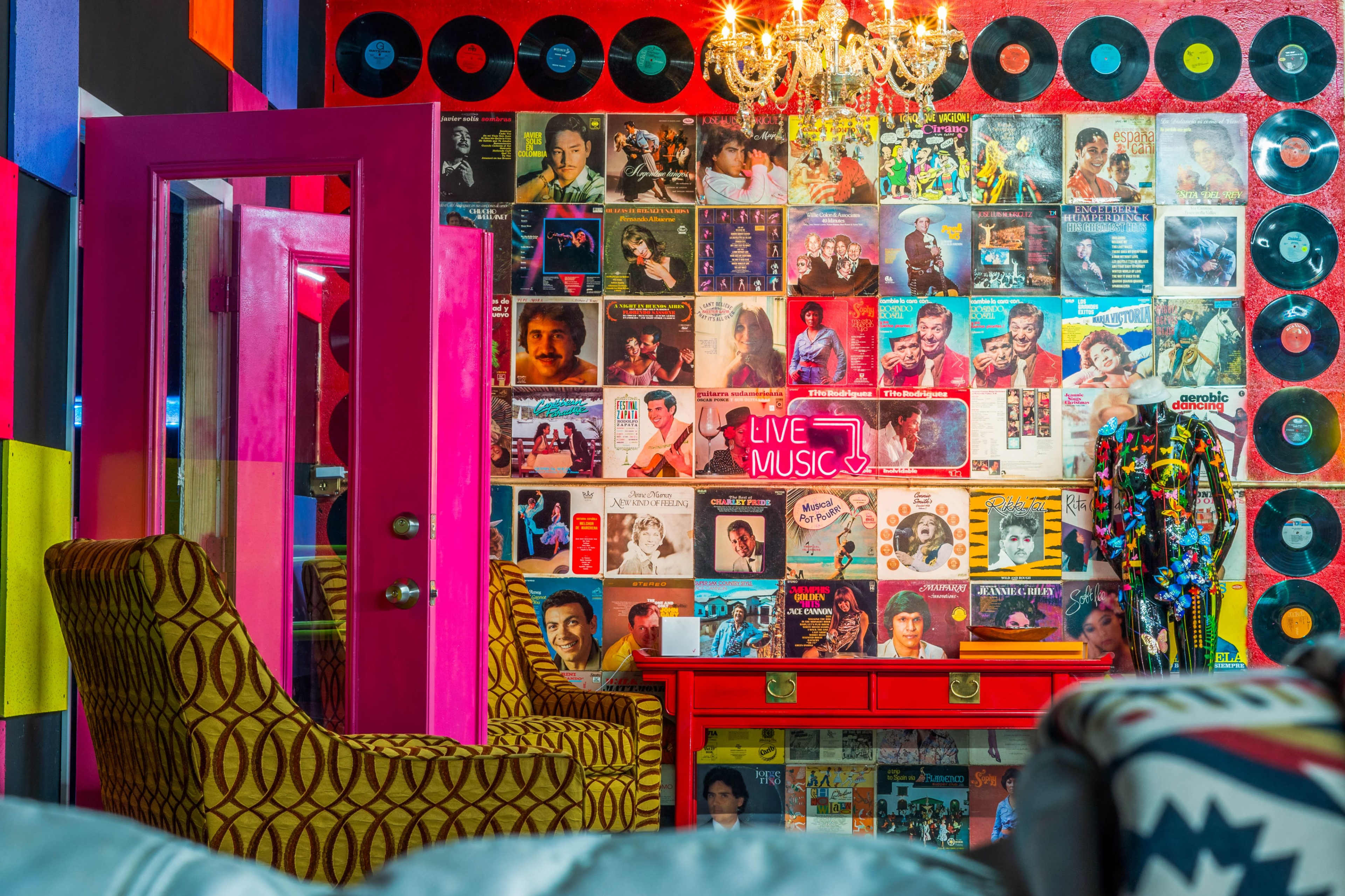 A vibrant room features a wall adorned with vintage vinyl records and movie posters, while a neon sign reading "LIVE MUSIC" adds a touch of illumination above a red cabinet and patterned chairs.