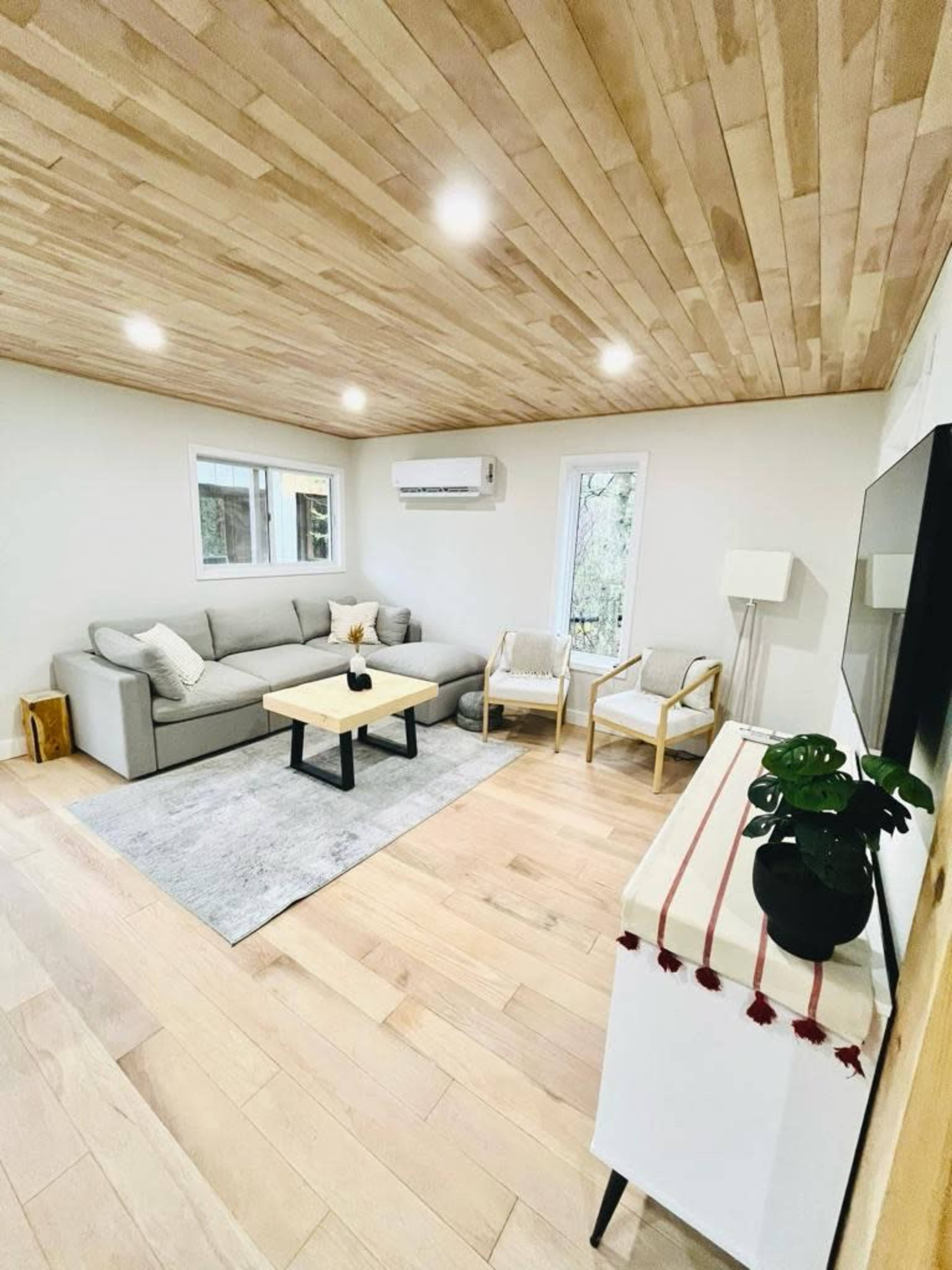 The image shows a cozy living room with a gray sofa, two chairs, a coffee table, and wood-paneled ceilings.