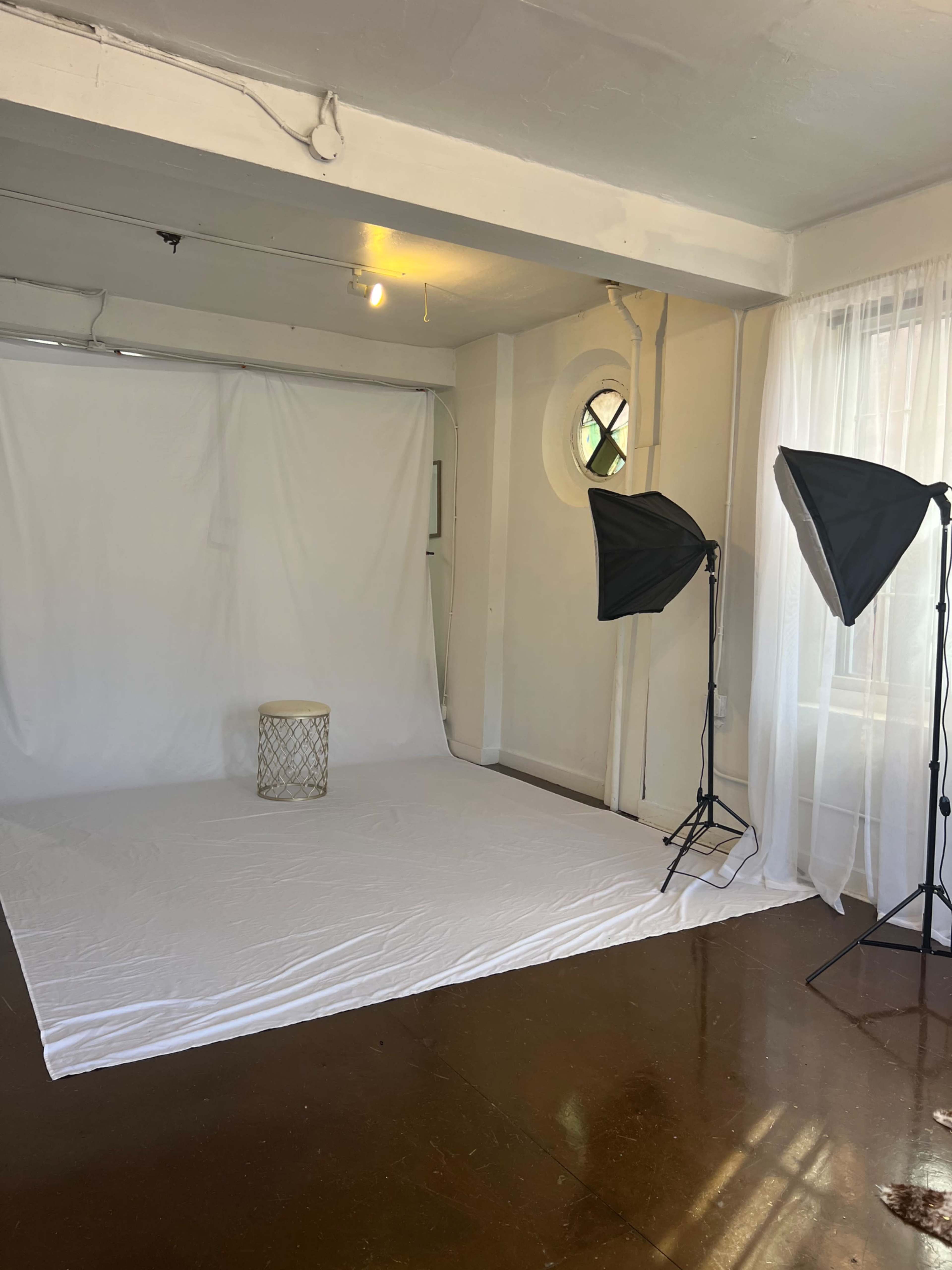 A photography studio with a white backdrop, a small round stool, and two lighting setup stands.