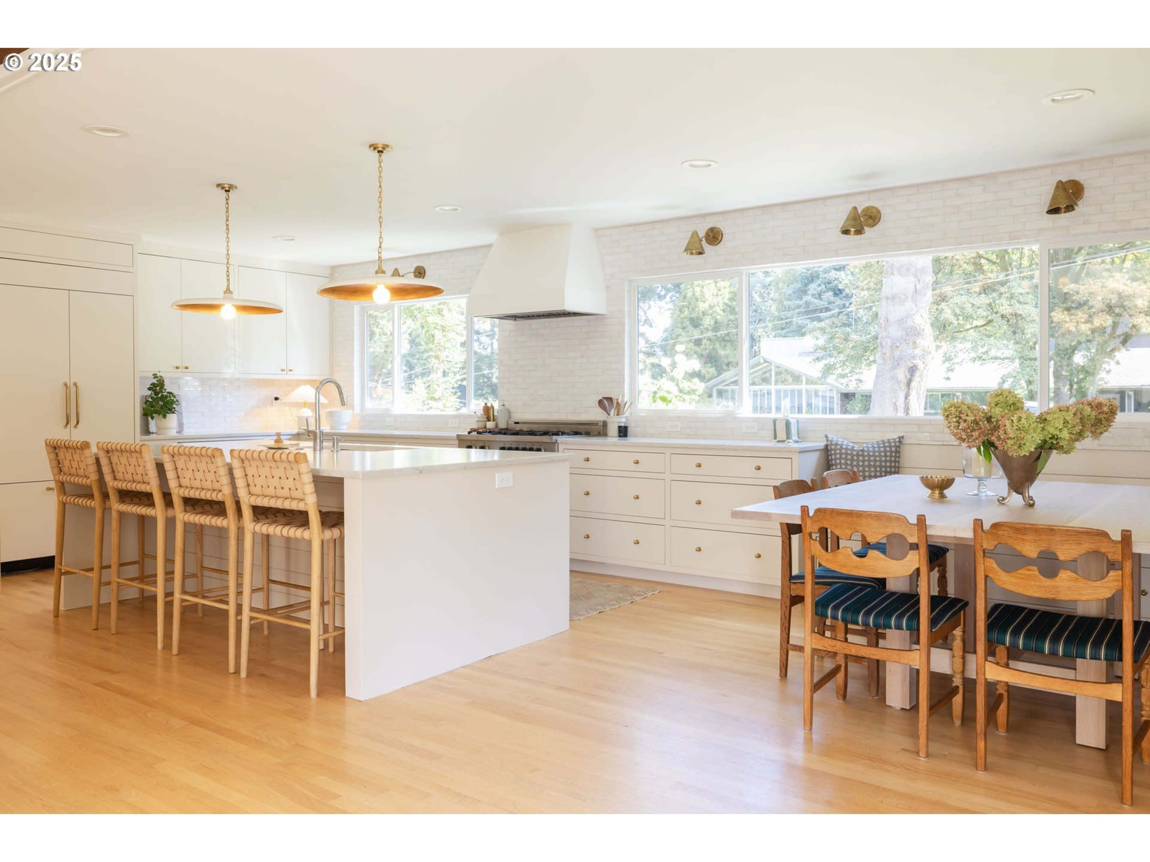 The image shows a modern kitchen featuring a large island with seating, white cabinetry, and a dining table next to a window that allows natural light to enter the space.