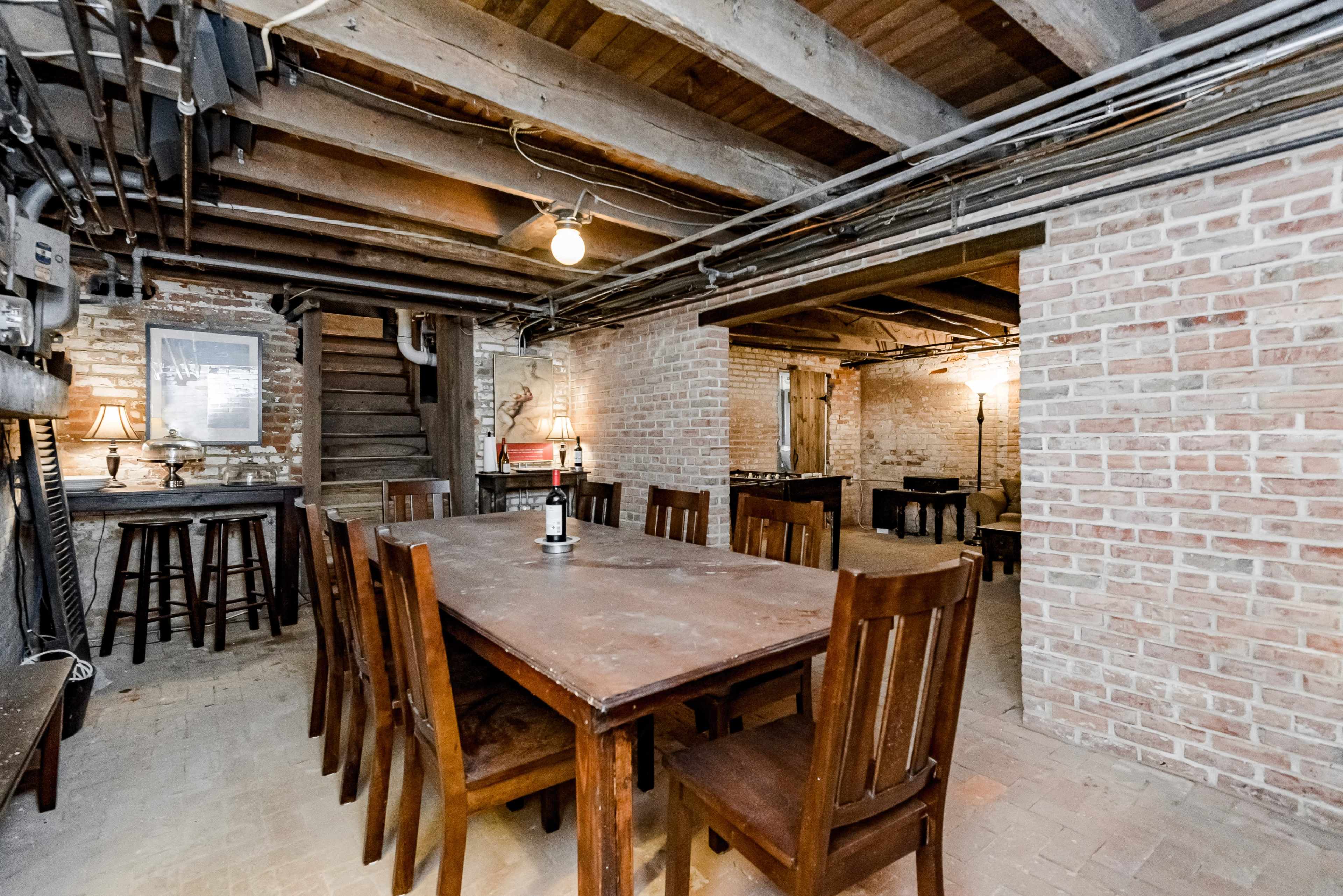 A spacious basement room with exposed brick walls, wooden beams, and a large wooden dining table surrounded by chairs.