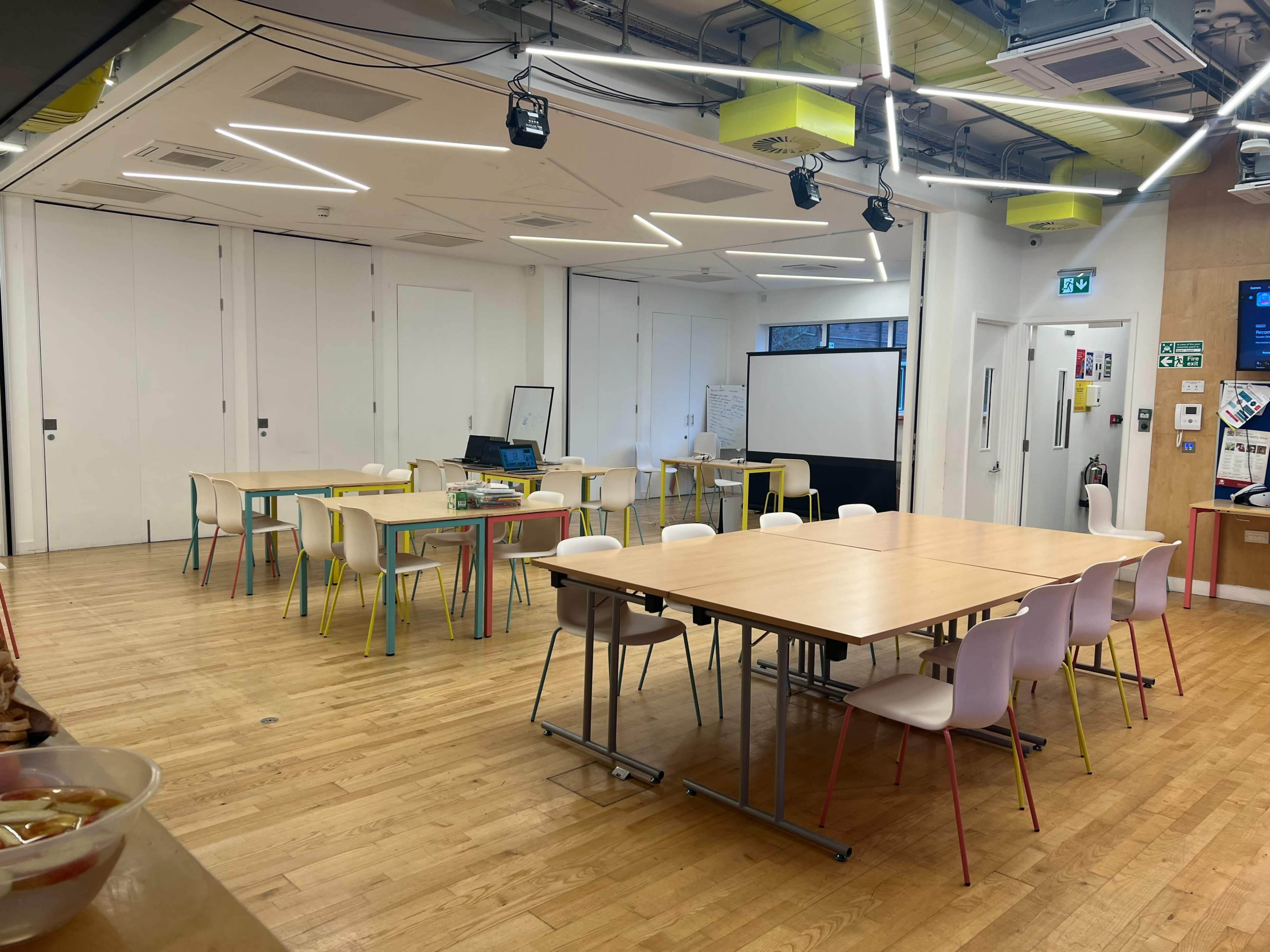 A modern classroom or meeting space with several wooden tables and white chairs arranged in a large open area.