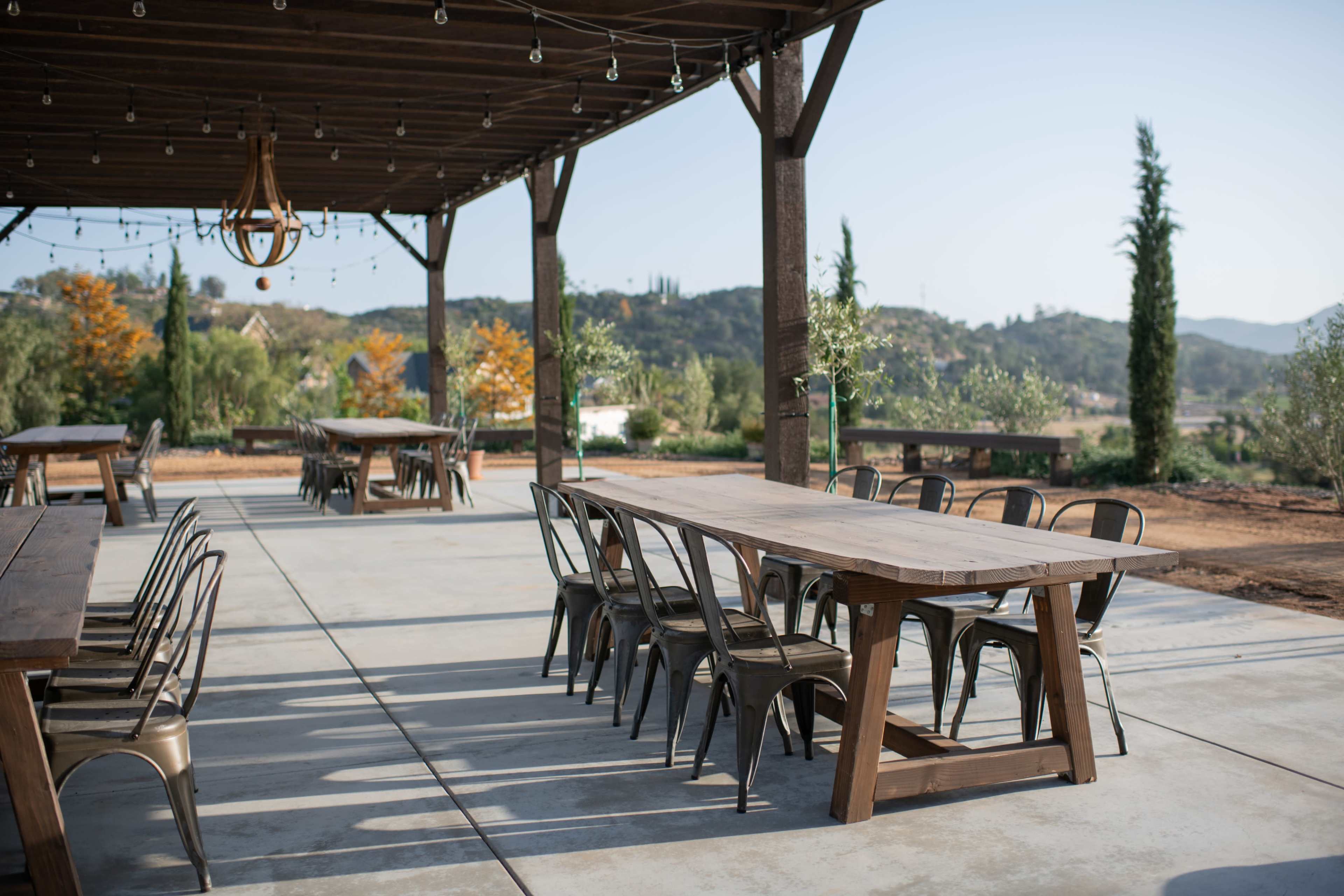 A spacious outdoor dining area features long wooden tables surrounded by metal chairs under a covered structure.