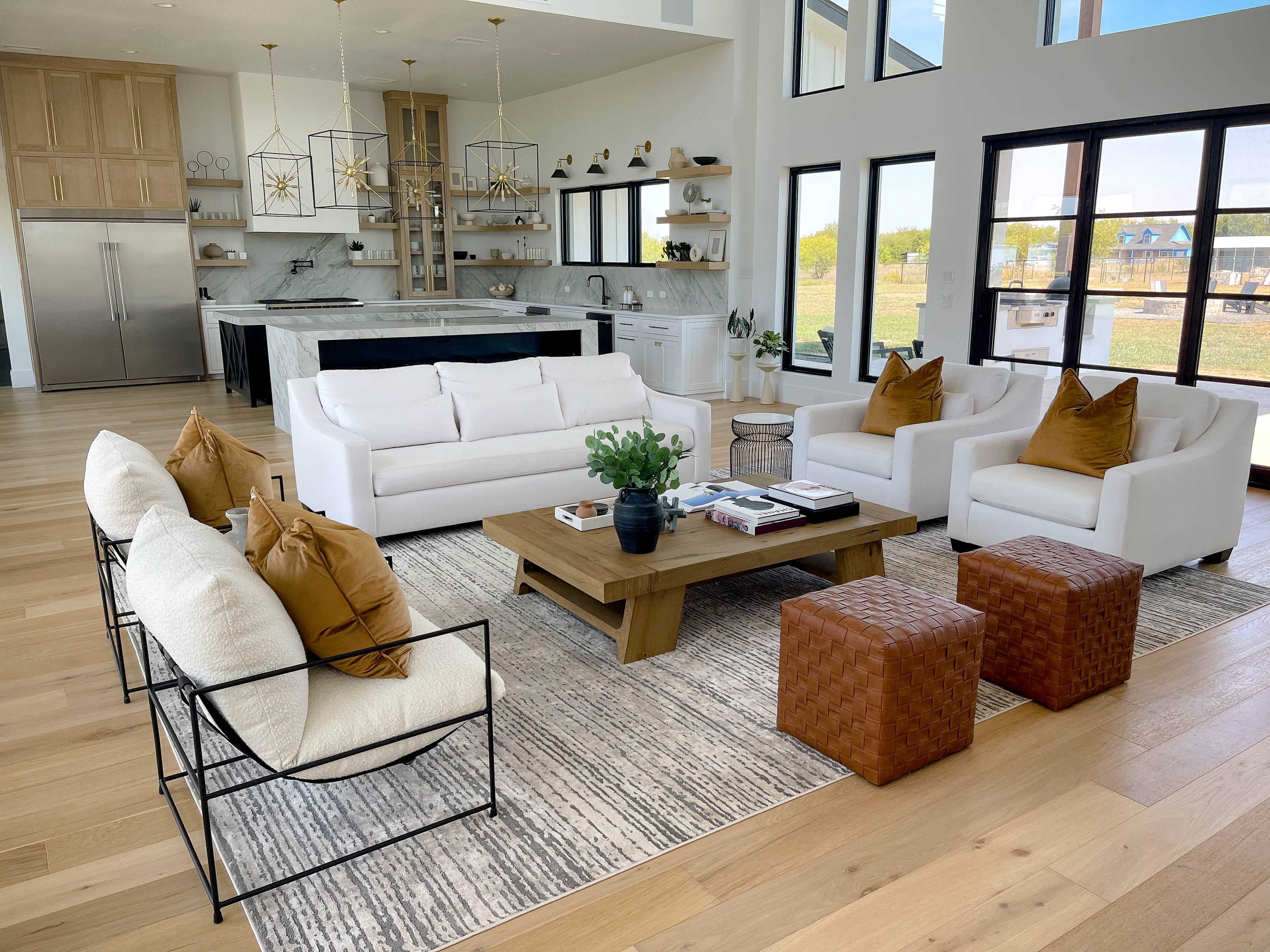 A modern living room features a large white sofa, two armchairs with decorative pillows, a wooden coffee table, and a patterned area rug, all arranged around a bright kitchen with large windows.