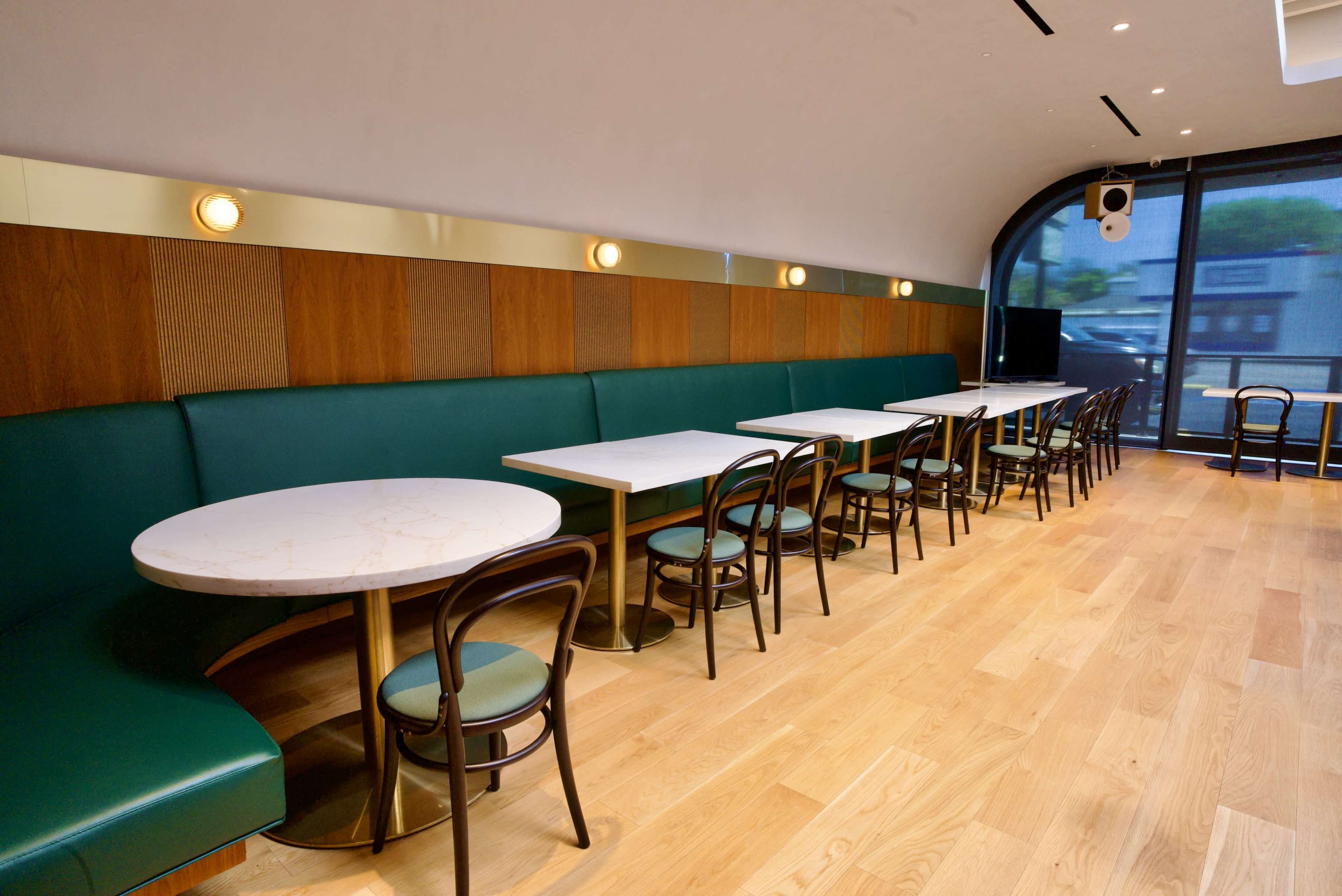 The image shows a modern café interior featuring a line of round tables with green chairs and a long green bench along a wooden wall.