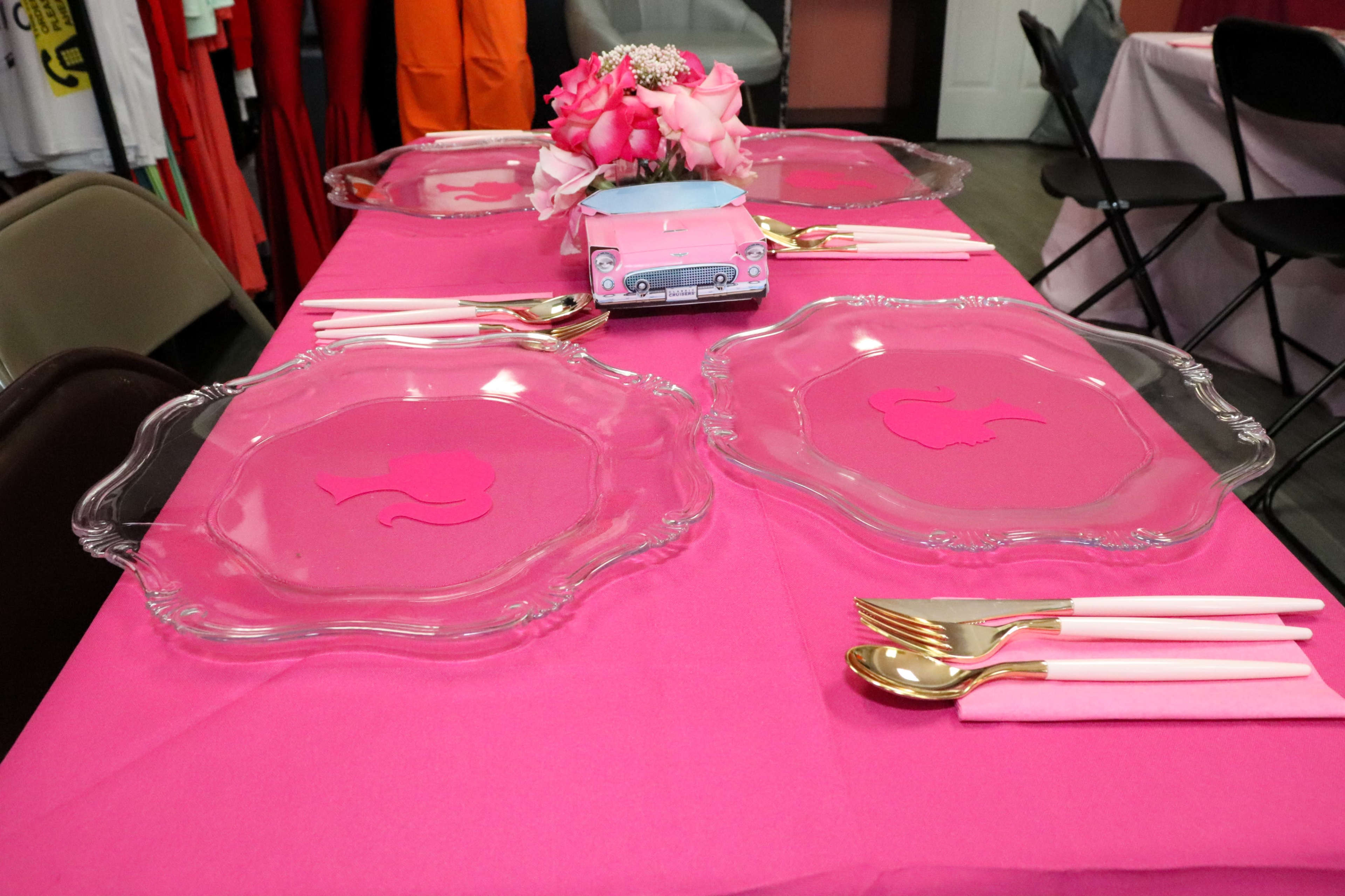 A pink table is set with glass plates and gold utensils, featuring a centerpiece of flowers and a decorative item.