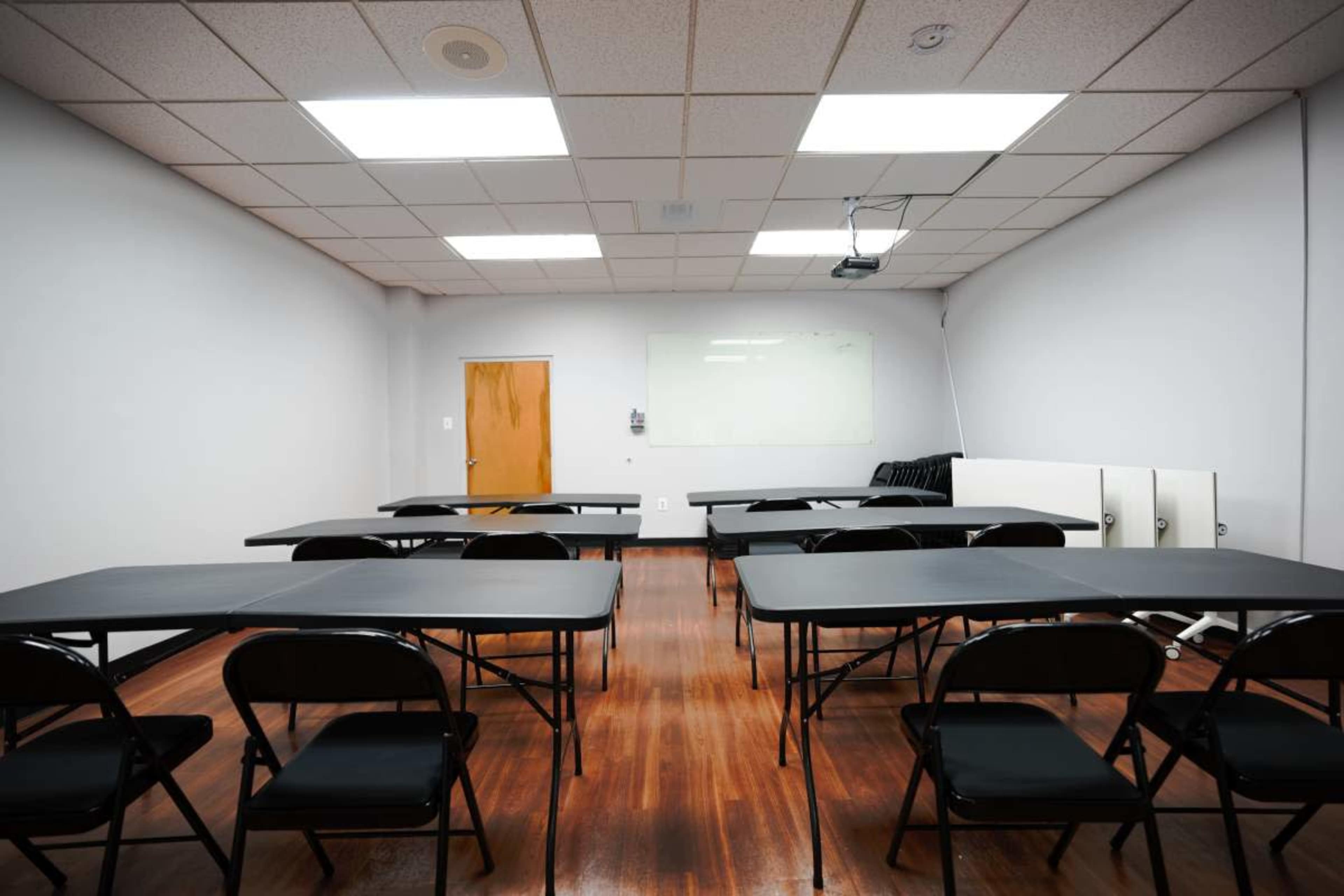 The image shows a classroom setup with several black tables and chairs arranged in rows, a whiteboard on the wall, and a projector mounted on the ceiling.