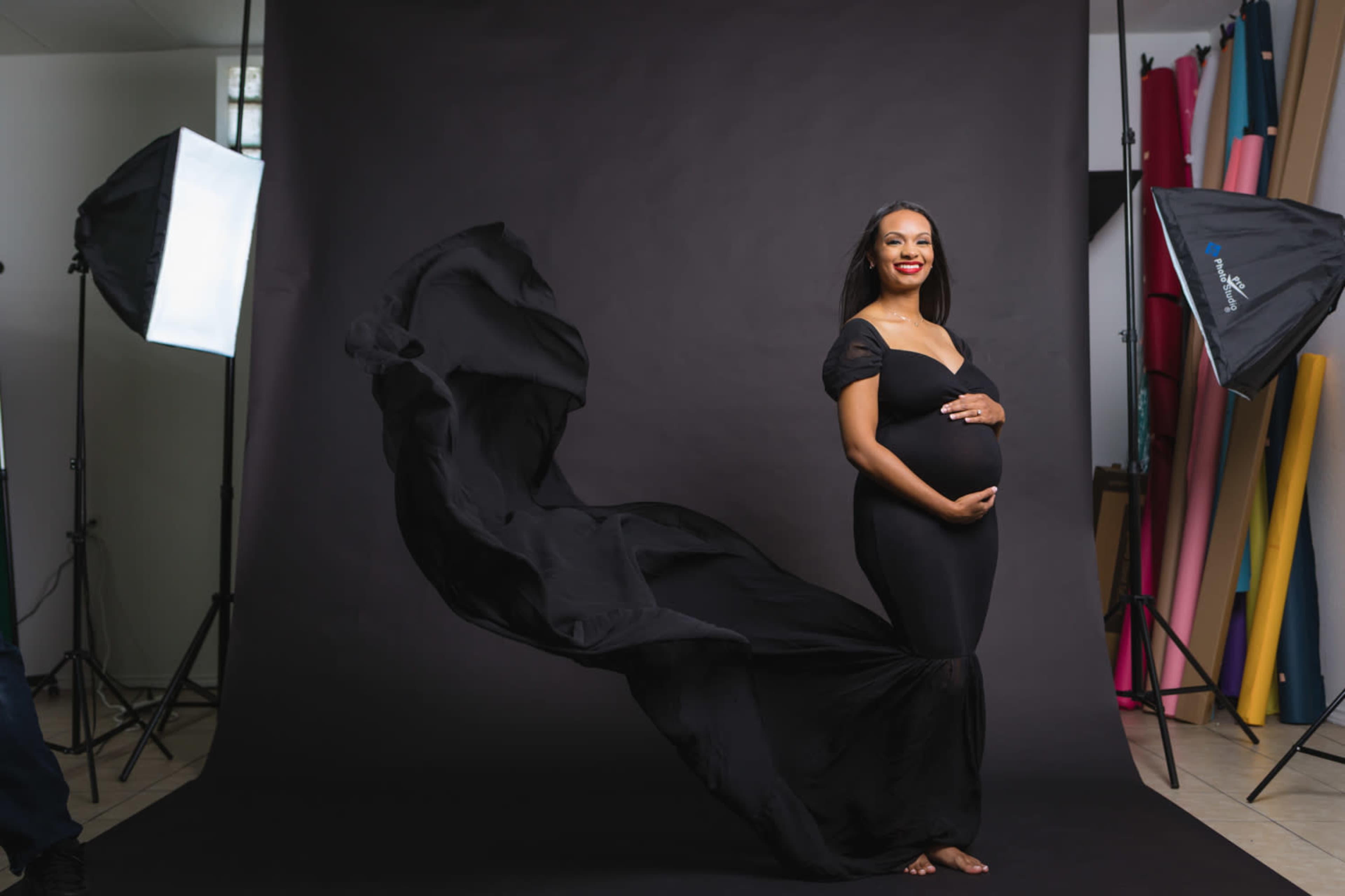 A pregnant woman stands confidently in a black gown against a dark studio backdrop, with fabric flowing around her and soft lighting illuminating the scene.