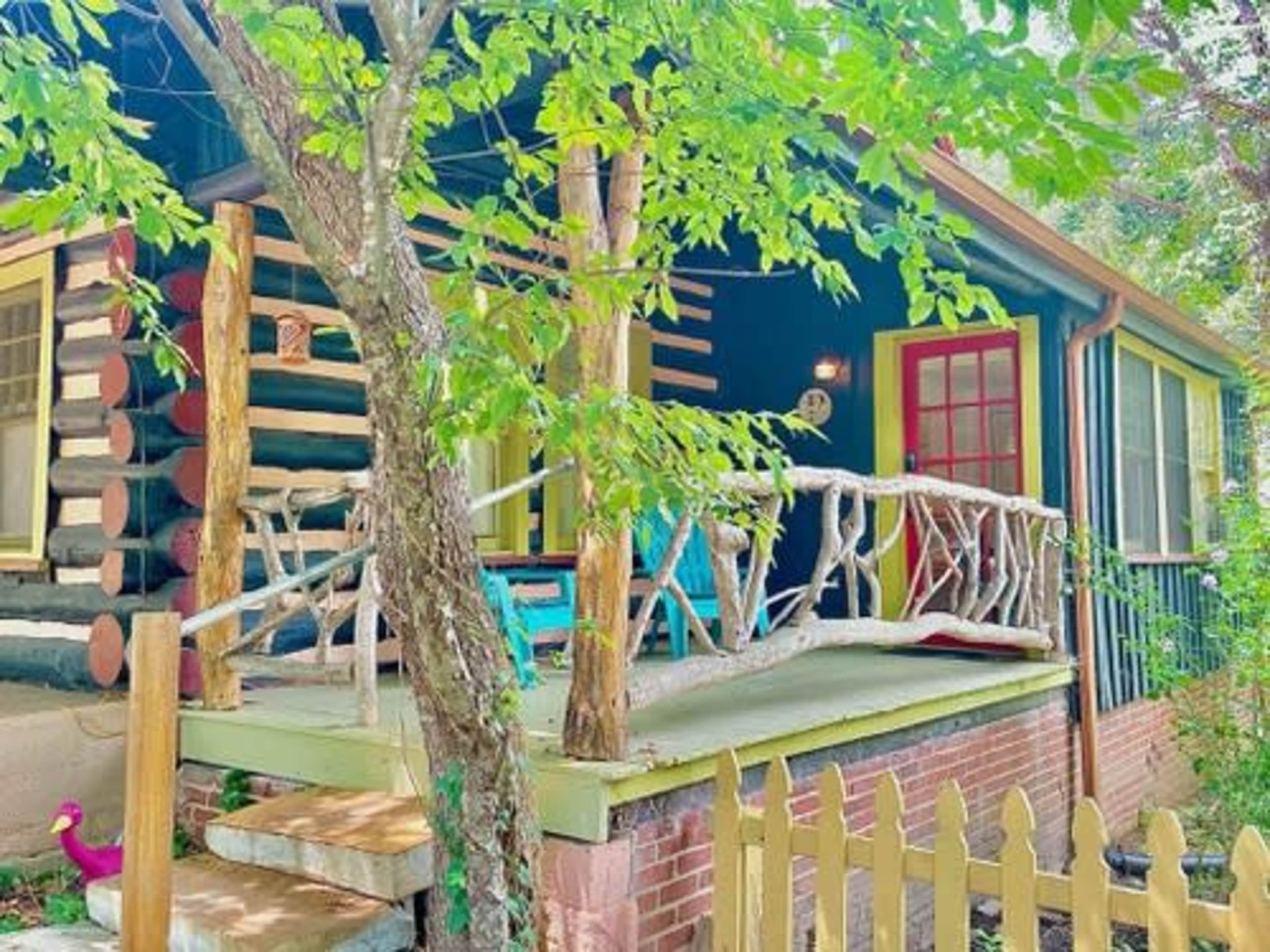 A small log cabin with a porch features a red door, colorful furniture, and a wooden railing surrounded by greenery.