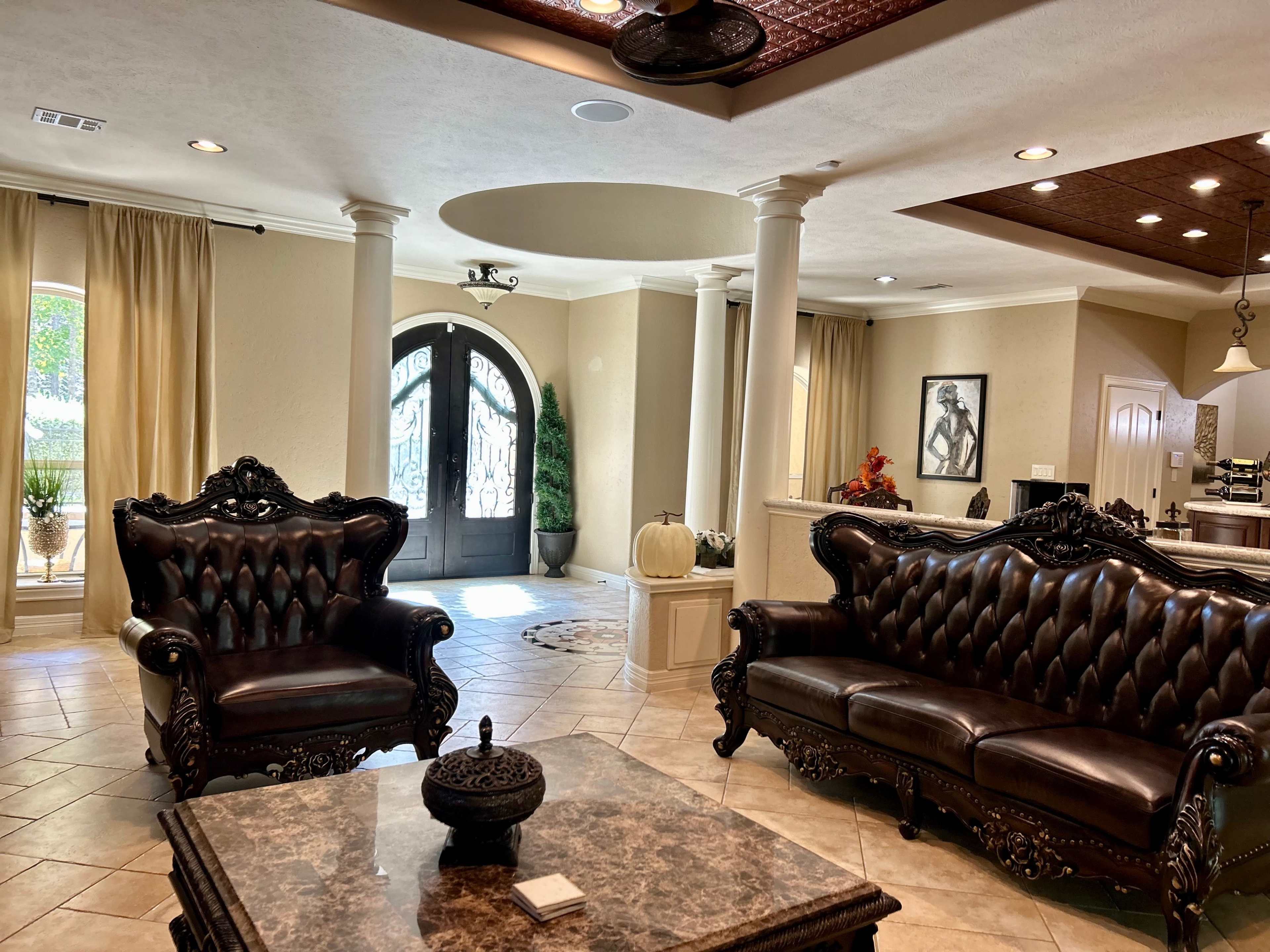 A spacious foyer features ornate black leather sofas, a marble coffee table, and a double-door entrance illuminated by natural light.
