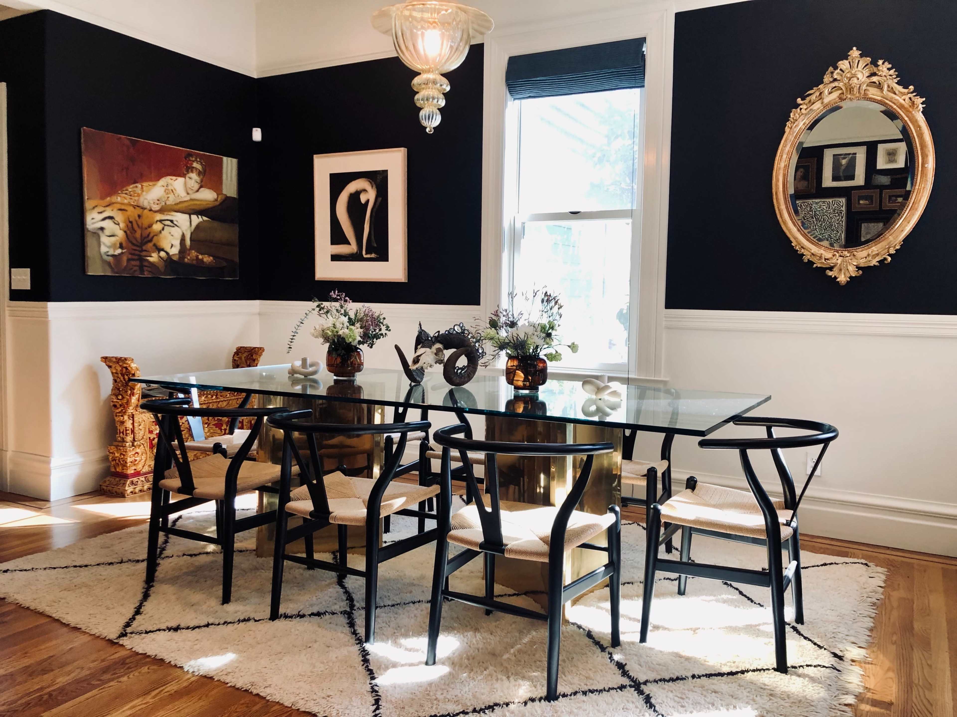 A dining room featuring a glass table surrounded by eight chairs, with a large window and black walls adorned with artwork and decorative mirrors.