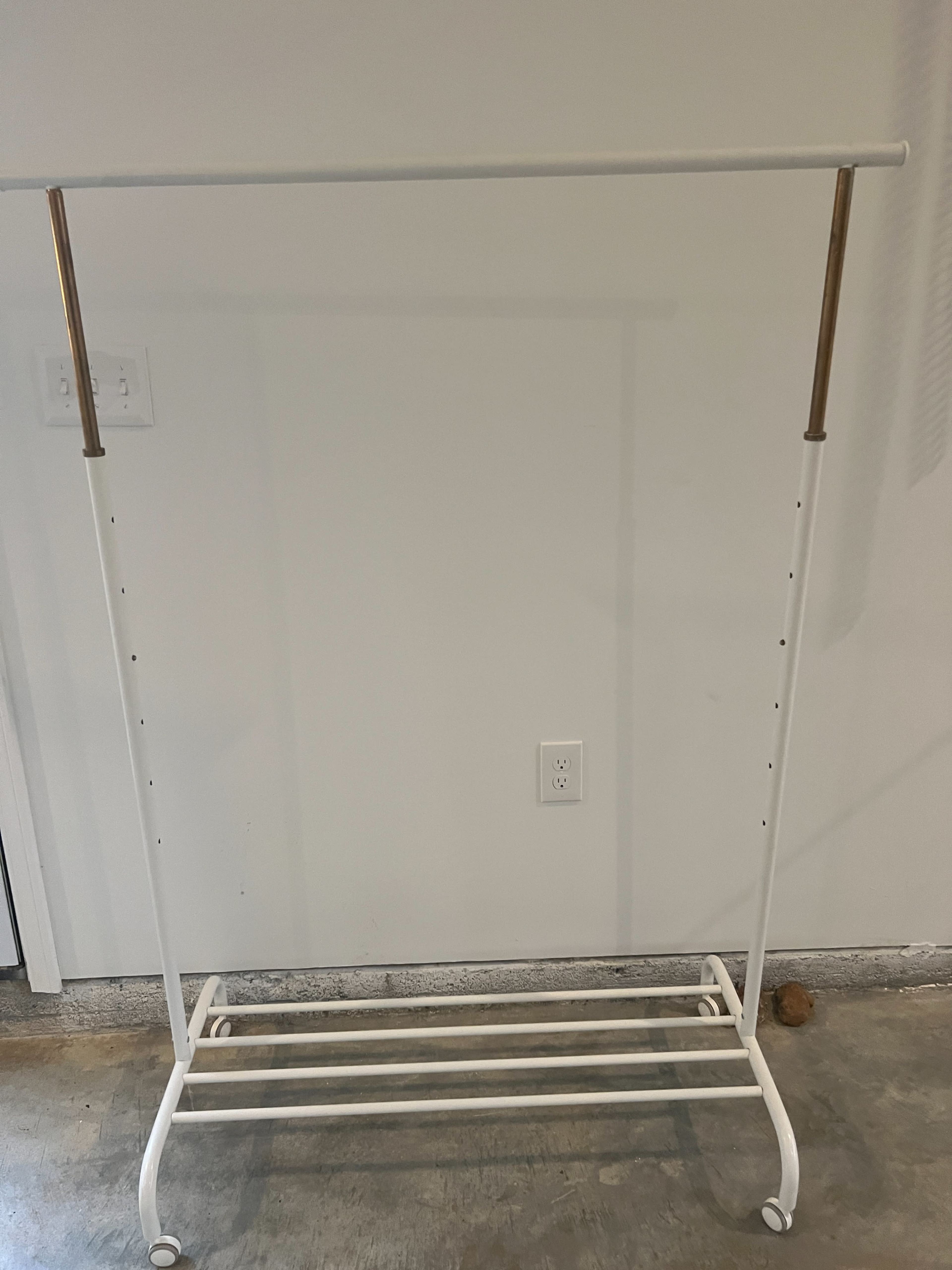 A white clothing rack with a horizontal bar and two gold-colored ends, sitting on a concrete floor.