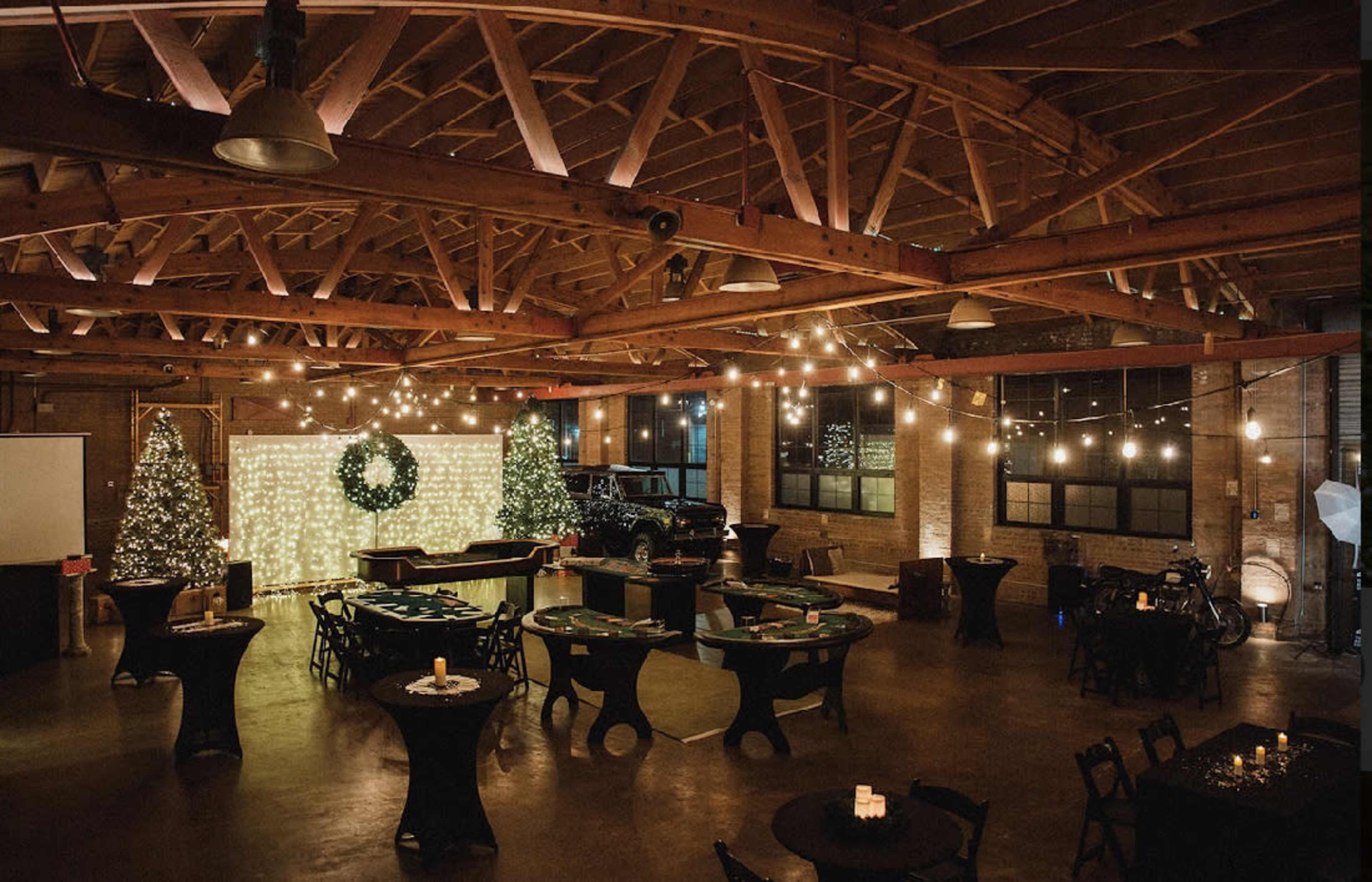 A spacious event venue with wooden beams, decorated with string lights and a festive backdrop, featuring tables arranged for a gathering.