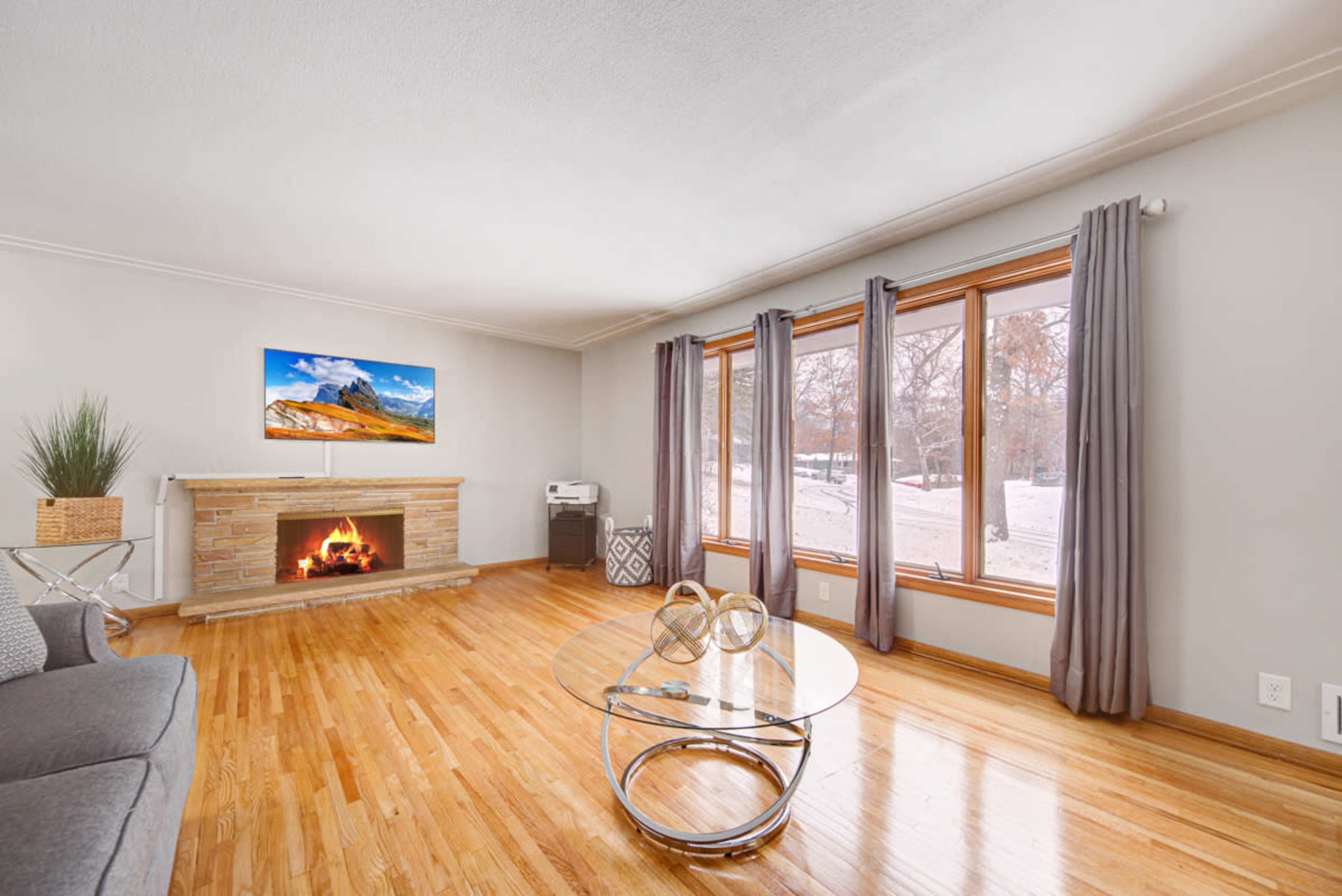 The living room features hardwood floors, a stone fireplace with a television above it, large windows with views of a snowy outdoor scene, and a round glass coffee table.