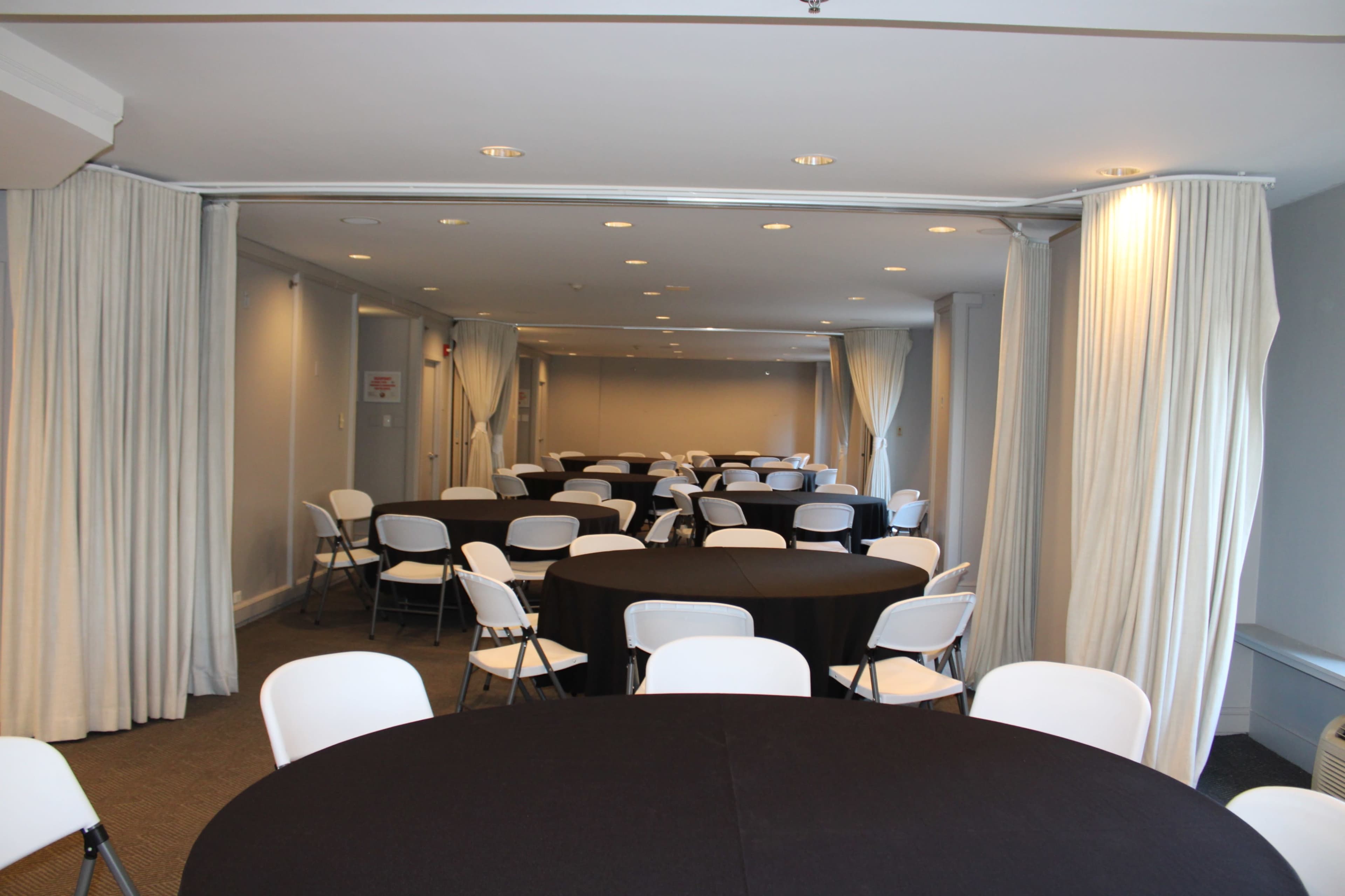 The image shows a meeting room with several round tables covered in black tablecloths and white chairs arranged throughout the space.