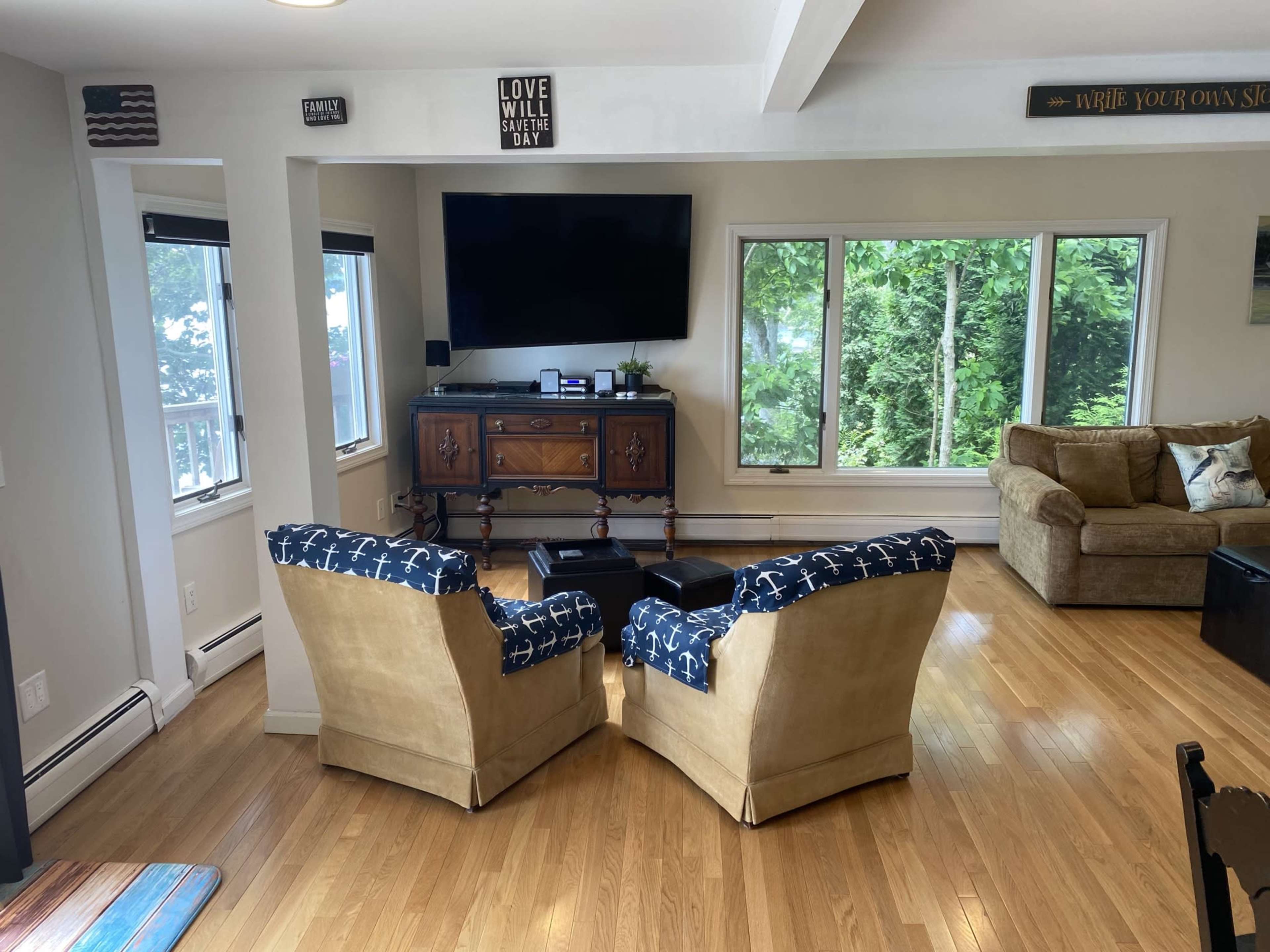 A living room features two beige chairs with blue patterned cushions facing a wall-mounted TV, surrounded by large windows offering a view of trees.