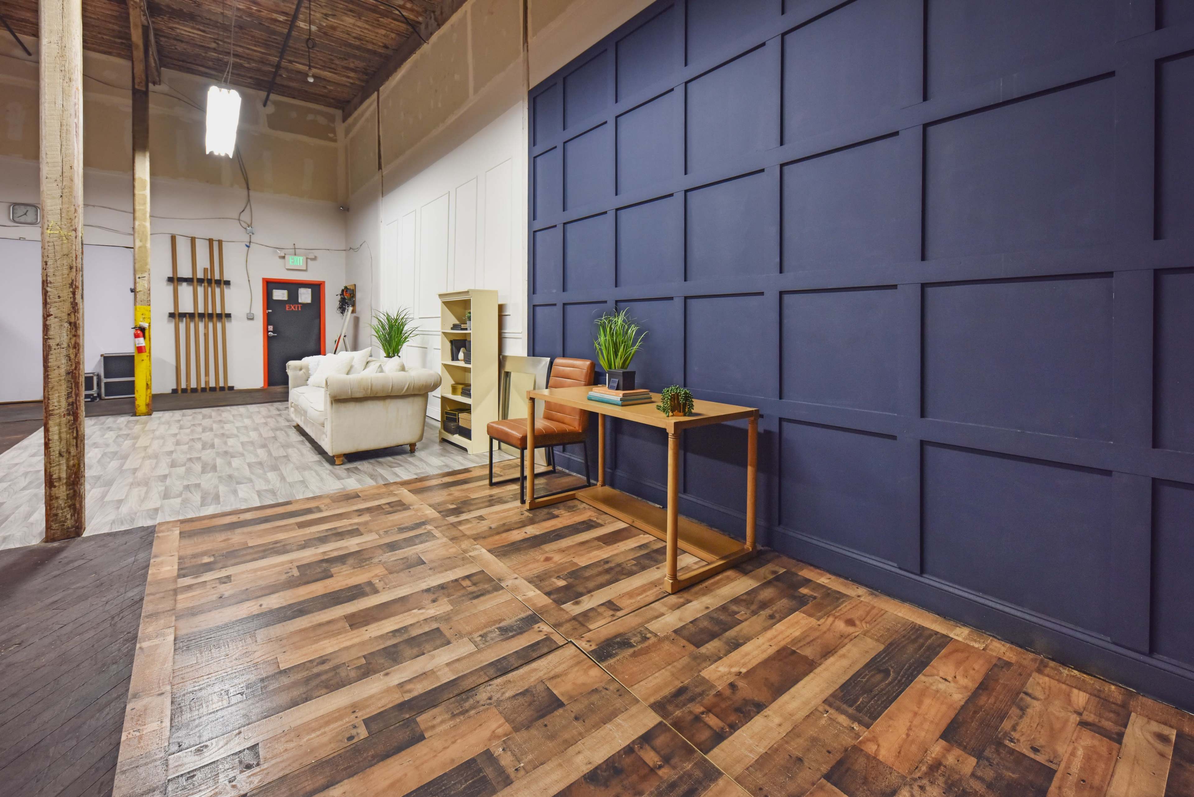 The image shows an interior space featuring a wooden floor, a dark blue accent wall, a simple desk with a plant on it, and a light-colored sofa in the background.