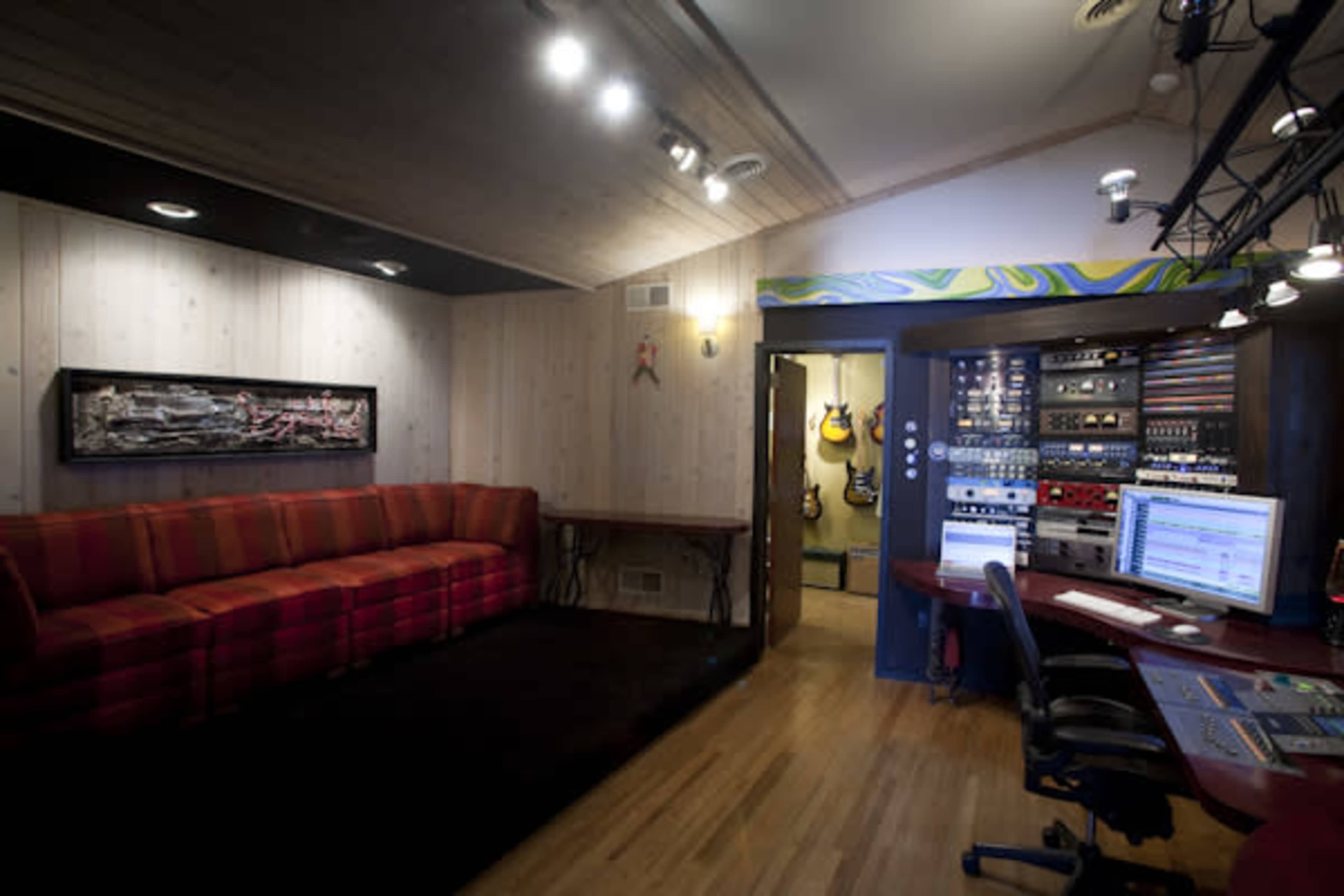 A sound studio featuring a red striped sofa, a workspace with computer monitors, and various musical instruments and equipment visible in the background.